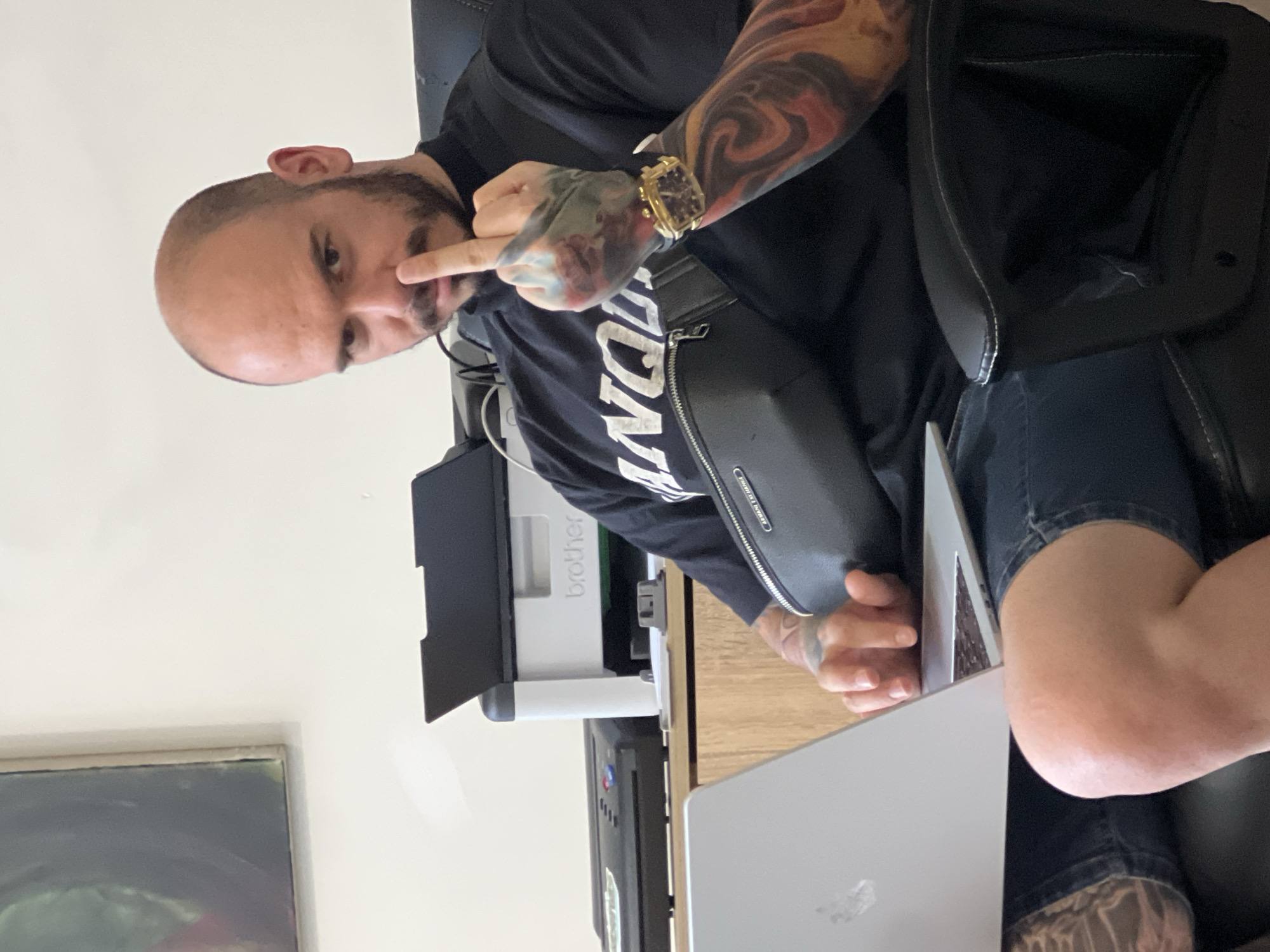 A man is seated indoors, wearing a black t-shirt, denim shorts, and a gold wristwatch. He has a shaved head, beard, and extensive tattoos on his arms. He is seated next to a table with a laptop and a Brother printer on it. The environment appears to be a home office. There is no evidence in the image to indicate this scene takes place in Sofia, Bulgaria.