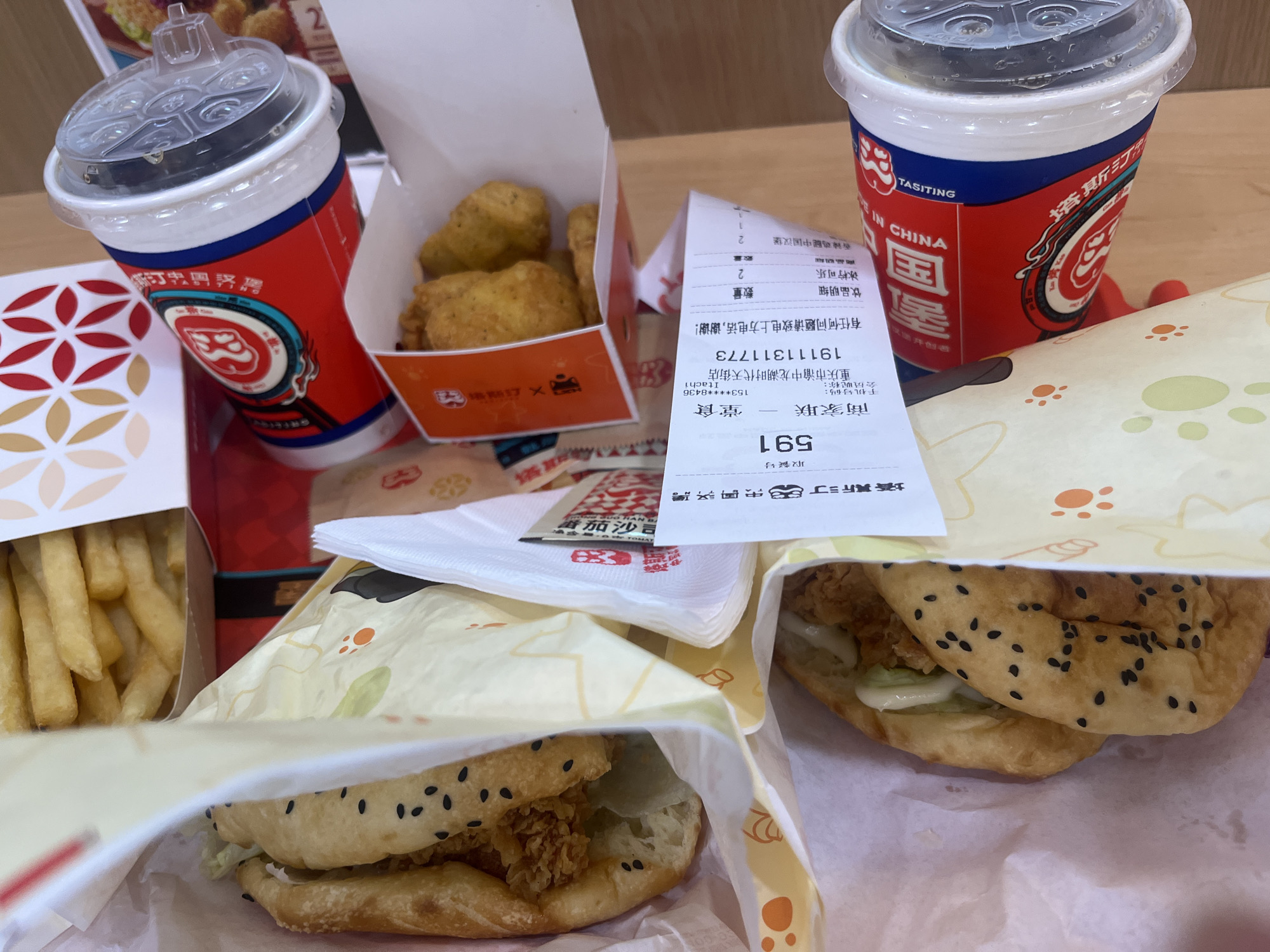 The image displays a close-up, top-down view of a fast-food meal spread out on a wooden surface, likely a table. The meal includes two drinks in red and white branded cups, one partially visible in the foreground left and another on the right. The cups feature Chinese characters and a logo. Adjacent to the drinks are boxes containing fried chicken nuggets and french fries, with the fries in a white and orange patterned container. A receipt is prominently placed in the center, showing Chinese text and numbers, including "1911131773" and "591". Two burgers, wrapped in patterned paper bags with orange paw prints, are positioned in the foreground, one on the left and one on the right, both revealing sesame seed buns and fillings. The overall scene suggests a casual dining experience, possibly a takeout order, in Yuzhong District, China, based on the receipt's inscription mentioning "重庆市渝中区".