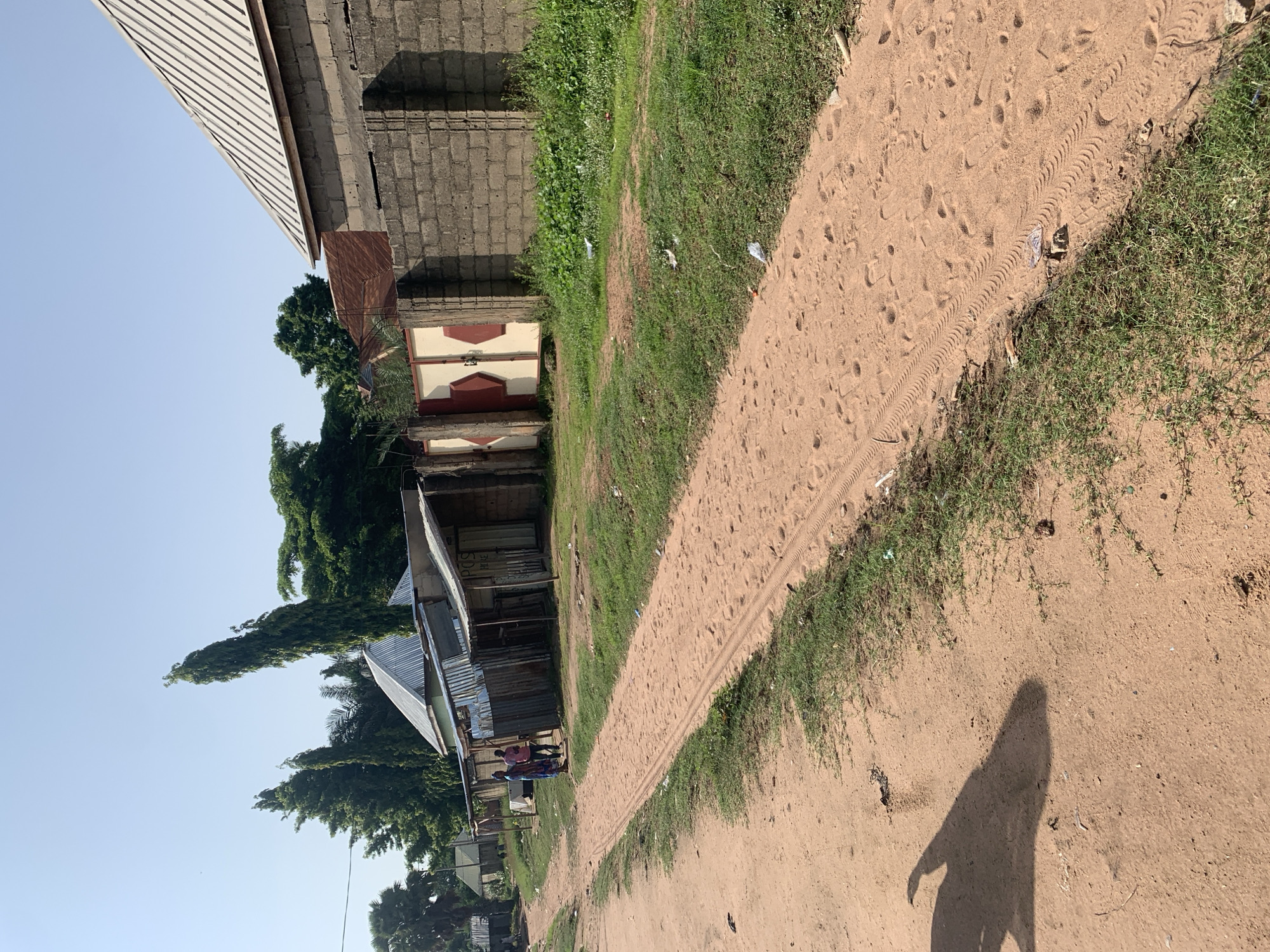 In Jalingo, Nigeria, a dirt road stretches across the frame, flanked by sparse vegetation and a few modest buildings. The road surface is sandy, marked by tire tracks and footprints. To the left, a shadow of an unseen object, possibly a person or a vehicle, is cast on the road. On the right side of the road, a low stone wall borders a property with a metal-roofed building and a large, partially visible stone structure. Further down the road, two individuals are visible standing near some roadside structures. The sky is clear and blue, indicating a sunny day.