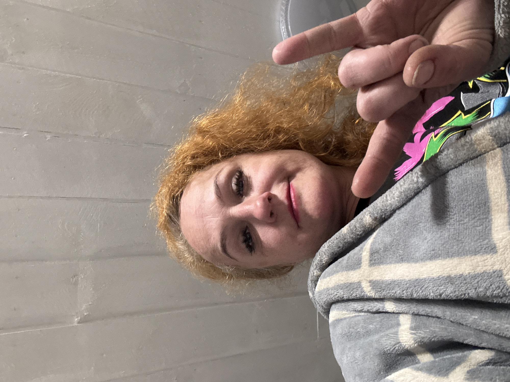 A woman with reddish-blonde curly hair is depicted from a low angle, slightly angled to the right. She is wearing a gray and white plaid robe. She is making a pointing gesture with two fingers of her right hand directed towards the lower part of her face. The background is a white wall with horizontal lines. The image was likely taken indoors, and the location within the city of Chaikovsky, Russia is not discernable from the visual information provided.