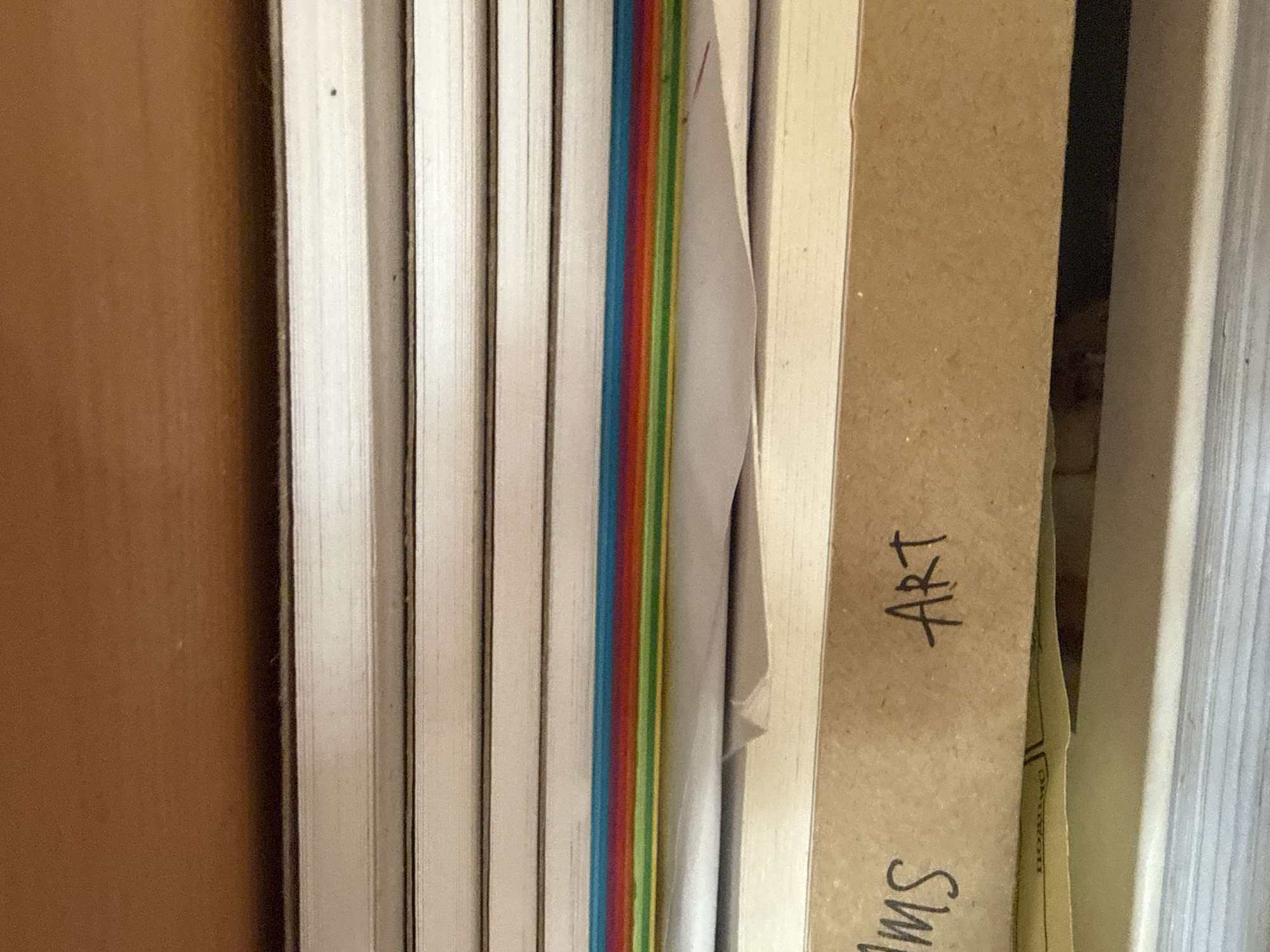 The image shows a close-up view of a stack of items in a drawer. Several items resemble books or notebooks with white spines. Between these, there is an object with multi-colored edges of blue, red, orange, and yellow. To the right of these items is a brown cardboard box with the word "ART" written on it. The drawer appears to be located indoors with a wood-paneled side. The exact location in the city of Troyan, Bulgaria is not discernible from the image.