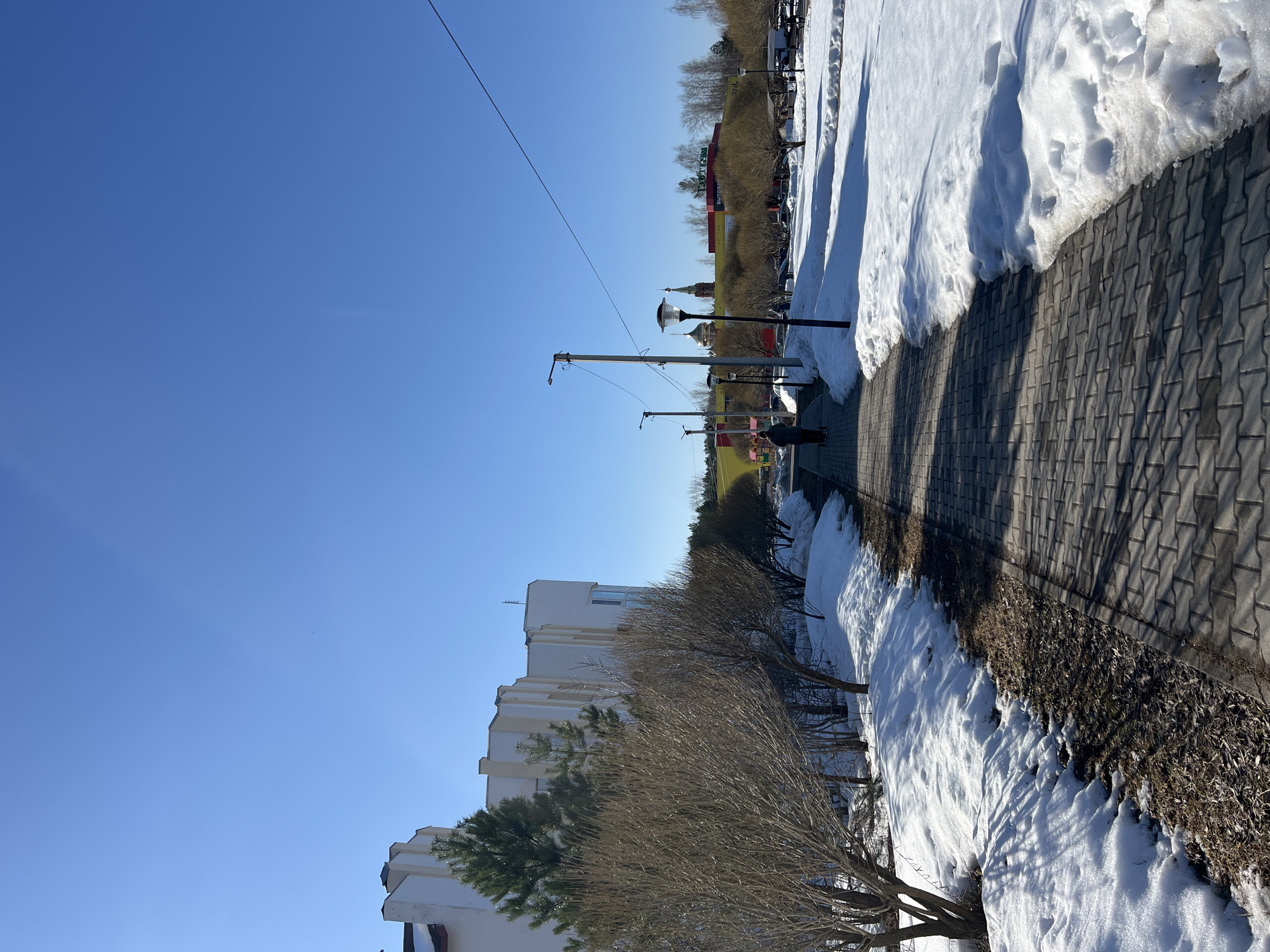 The image depicts a winter scene in Chaikovsky, Russia. A paved walkway, constructed from gray interlocking paving stones, runs through the scene. Patches of snow line the path. Trees and shrubs are planted alongside the walkway. In the background, there is a building with white walls. Further back, a tower with a red roof is visible. Overhead power lines stretch across the clear blue sky. There are also lamp posts along the path.