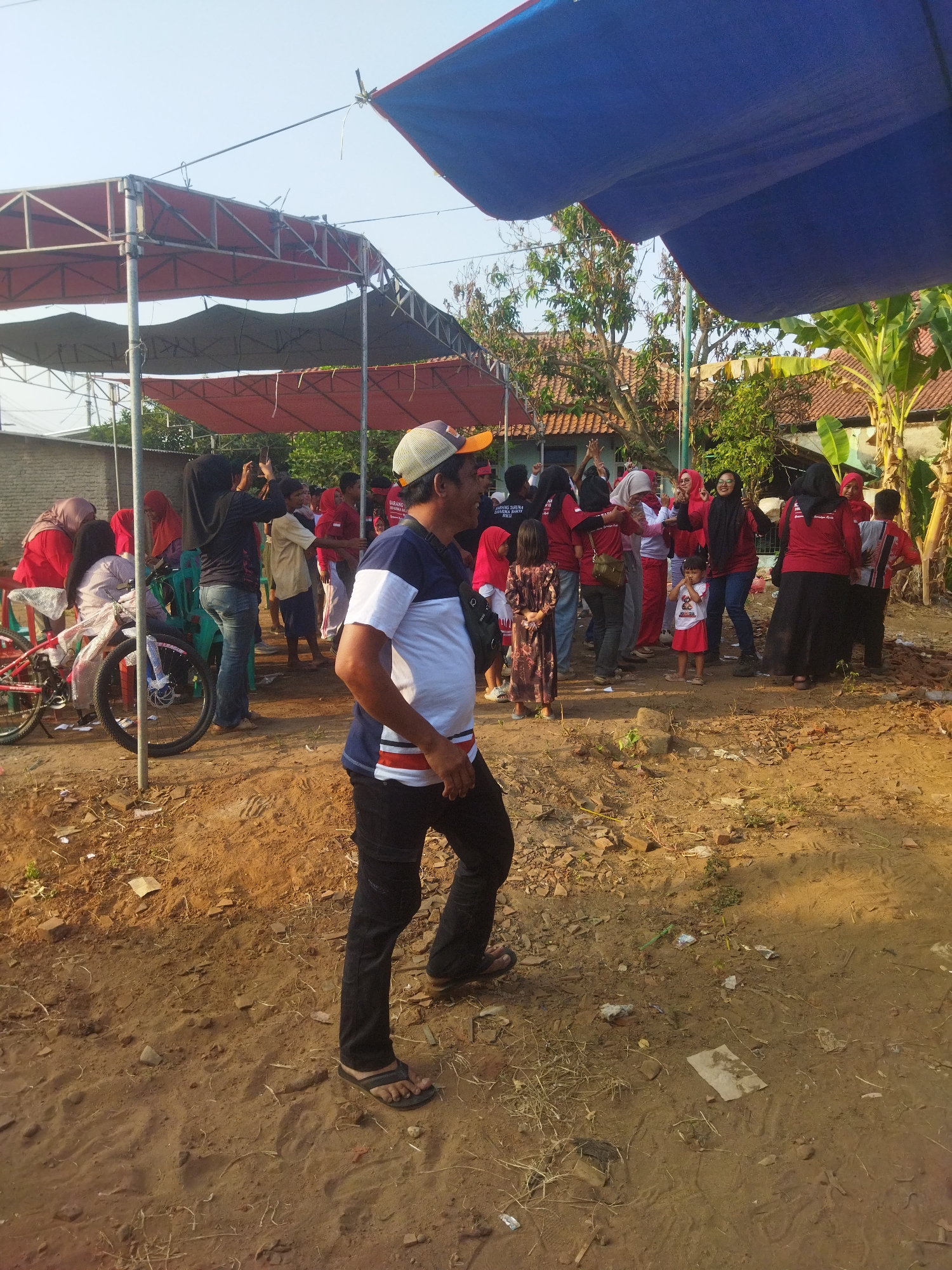 The image depicts an outdoor gathering in what appears to be a residential area in Banjar, Indonesia, likely during daytime given the bright lighting. In the foreground, a man wearing a blue and white striped shirt, black pants, and a baseball cap is walking towards the right of the frame. He is casually dressed, with dark sandals on his feet and a black bag slung across his chest.
Behind him, a group of people are congregated under makeshift tents. The tents are supported by metal poles and covered with red and blue tarpaulins, suggesting an event or temporary structure for shade. Many individuals, including women and children, are seen wearing bright red clothing, possibly indicating a team or celebratory attire. Some are interacting, with some women wearing headscarves. A red bicycle with a woman seated on it is visible to the left. The ground is primarily dirt and sand, with some scattered debris, indicating an undeveloped or natural outdoor space. The background shows houses with tiled roofs and lush green vegetation, typical of a tropical environment. The overall scene suggests a community event, possibly a festival or social gathering.