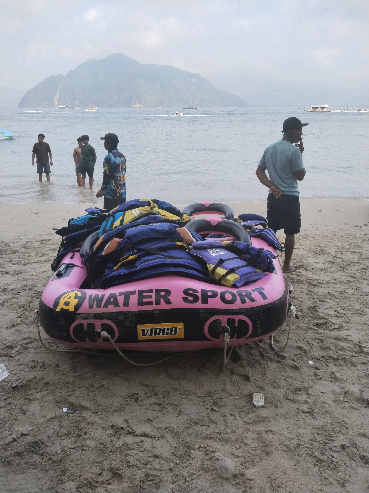 On a sandy beach, a bright pink inflatable raft sits partially submerged in shallow water. The raft is emblazoned with the words "WATER SPORT" in black lettering and the brand name "VIRGO". It is laden with numerous blue and yellow life jackets, piled high. Four men are visible. Three stand in the water near the shore, their legs submerged. One man, wearing a dark t-shirt and shorts, is facing away from the camera. Another, in a green t-shirt and dark shorts, stands beside him. The third, in a dark, patterned long-sleeved shirt and dark shorts, faces towards the raft. A fourth man, wearing a light blue t-shirt and black shorts, stands on the dry sand to the right of the raft, with his back to the camera, holding what appears to be a cigarette. In the background, the calm sea stretches to a hazy horizon, with a large, mist-covered mountain dominating the landscape. Several boats and structures are visible on the water, including what appears to be a floating platform. The scene suggests a recreational area, possibly the beach at Prigi, Indonesia, known for its watersports activities.