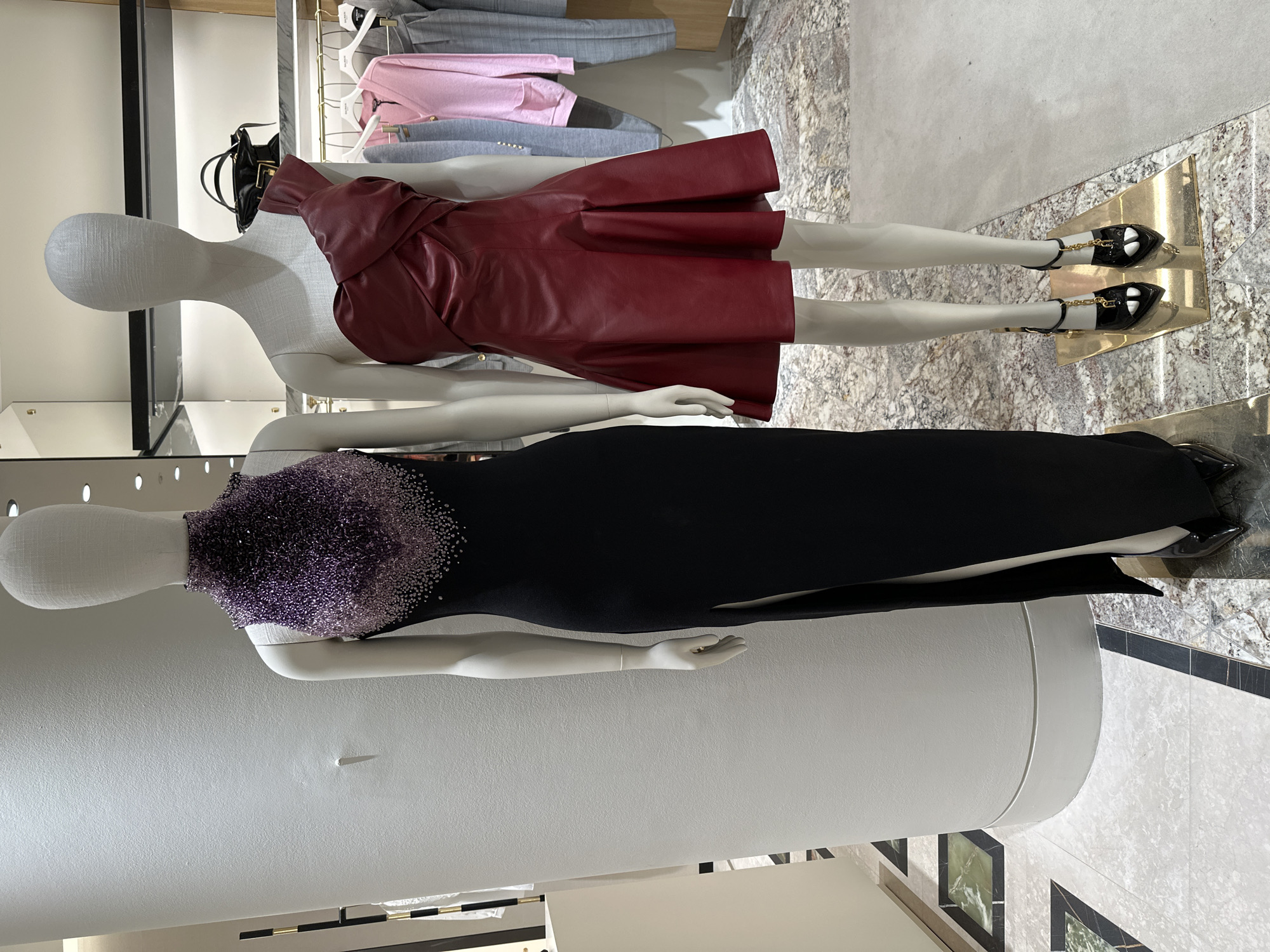 The image shows two mannequins on display inside a shop in London, United Kingdom. The upper mannequin is wearing a red leather dress and black shoes with gold chain accents. A black handbag sits on the shelf behind the mannequin. The lower mannequin wears a black dress with purple beadwork around the collar. The flooring is marble patterned. Garments are hanging on a rail in the background.