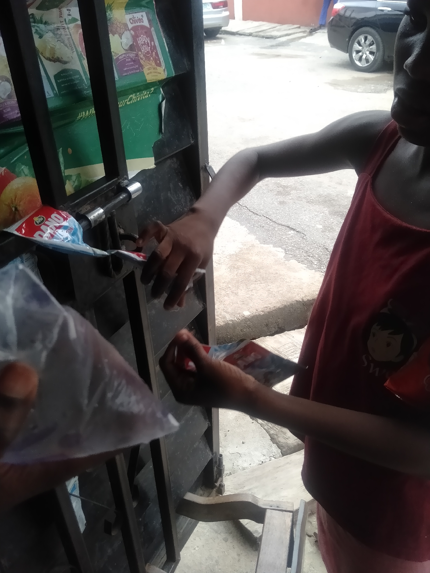 This image captures a street vendor transaction in an outdoor setting, likely in Shangisha, Nigeria.
**Subjects:** A person, possibly a youth, is visible on the right side of the frame, wearing a dark red sleeveless top with a cartoon graphic and the word "SWEET" printed on it. Their face is largely obscured by shadow. Another person's hand is seen in the lower left, holding a clear plastic pouch.
**Objects:** The central object is a dark metal vendor stall or kiosk, constructed with bars. Inside the stall, several packaged products are displayed. Prominently visible are "Chivita Happy Hour" juice pouches, with text mentioning "PINEAPPLE & COCONUT" and "TASTY MANGO" flavors. A small "Dano" sachet, likely milk powder, is also on the stall surface. The person on the right is holding a small red and white sachet, while the hand on the left holds a clear plastic pouch filled with a dark liquid or substance. In the background, a black sedan car is parked on a concrete surface. A wooden stool or bench is partially visible at the bottom.
**Activity:** The image depicts an exchange between the two individuals, suggesting a purchase or sale at the vendor stall. The person on the right appears to be handling goods, while the person on the left holds a product in a plastic pouch.
**Notable Details:** The scene takes place during the daytime, possibly in a shaded area or under an overcast sky, given the soft lighting and deep shadows. The use of clear plastic pouches for beverages or snacks is a common sight with street vendors in regions like Nigeria. The specific brands "Chivita" and "Dano" are well-known in the Nigerian market.