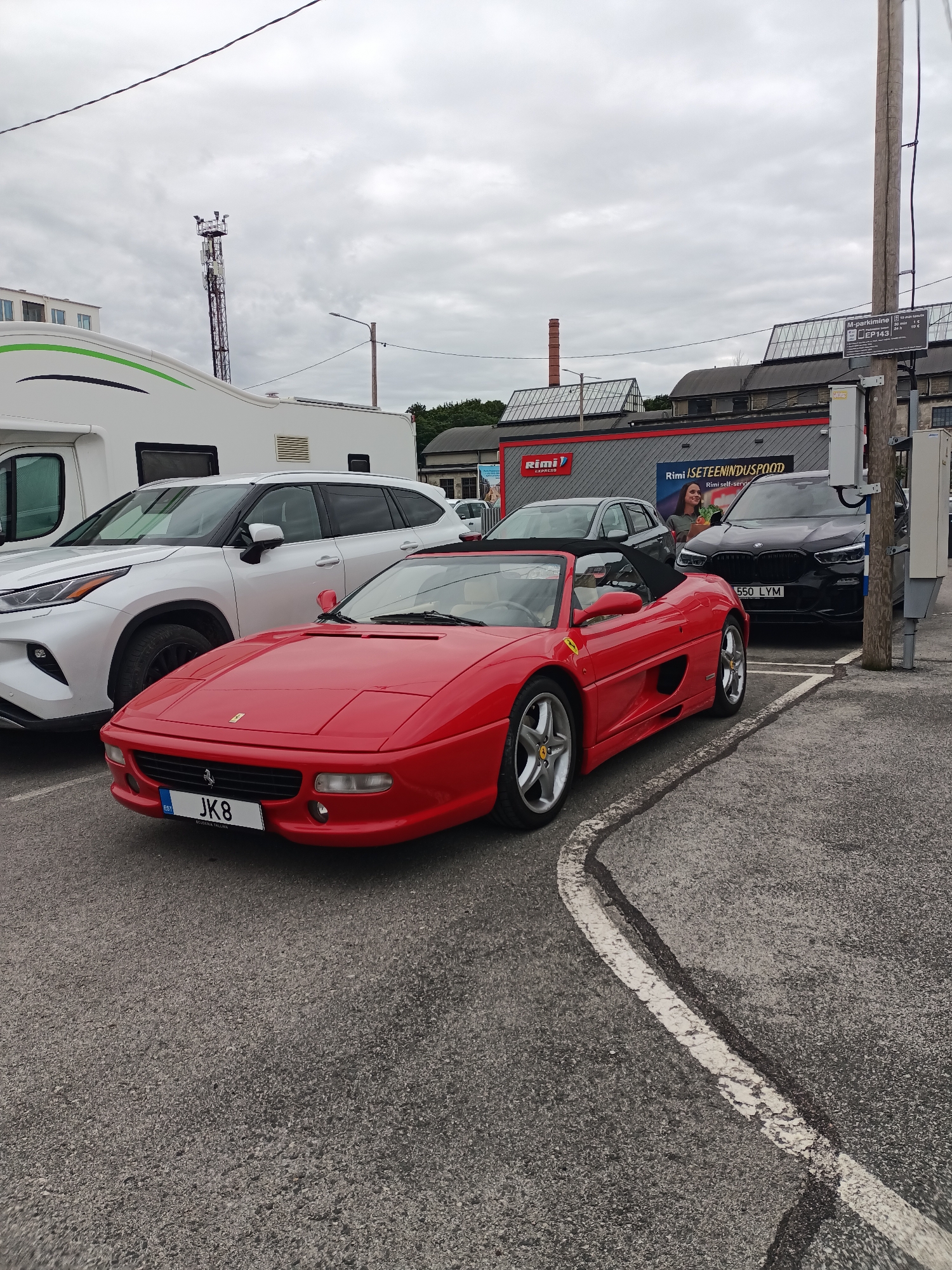 A red Ferrari convertible is parked on asphalt in a lot with white painted lines. To the left of the Ferrari is a white SUV, and to the left of the SUV is a white RV. In the background, there is a dark SUV, a building with a red sign that says "Rimi", and a telephone pole with a parking meter. The sky is cloudy. The license plate on the Ferrari is "JK8". The license plate on the dark SUV is "550 LYM". The image was taken in Tallinn, Estonia.