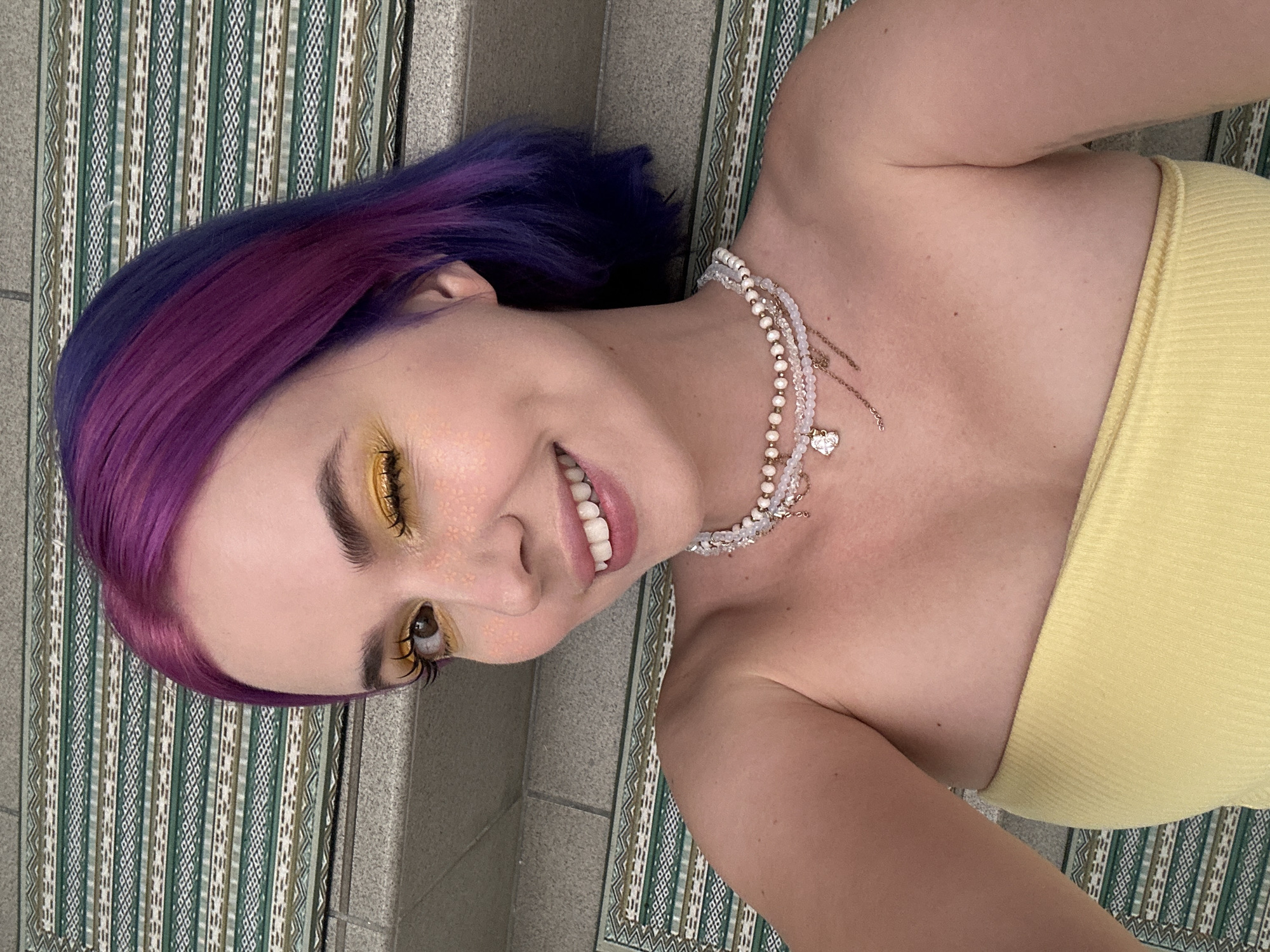 A person lies on a patterned rug placed atop a concrete floor, smiling towards the camera. The individual has hair that transitions from purple to magenta and is wearing yellow eyeshadow. A multi-strand pearl necklace adorns their neck, and a yellow strapless top is visible. The rug has a repeating geometric pattern in shades of green, white, and brown. The concrete floor, likely within City of Zagreb, Croatia, is visible beneath the rug.