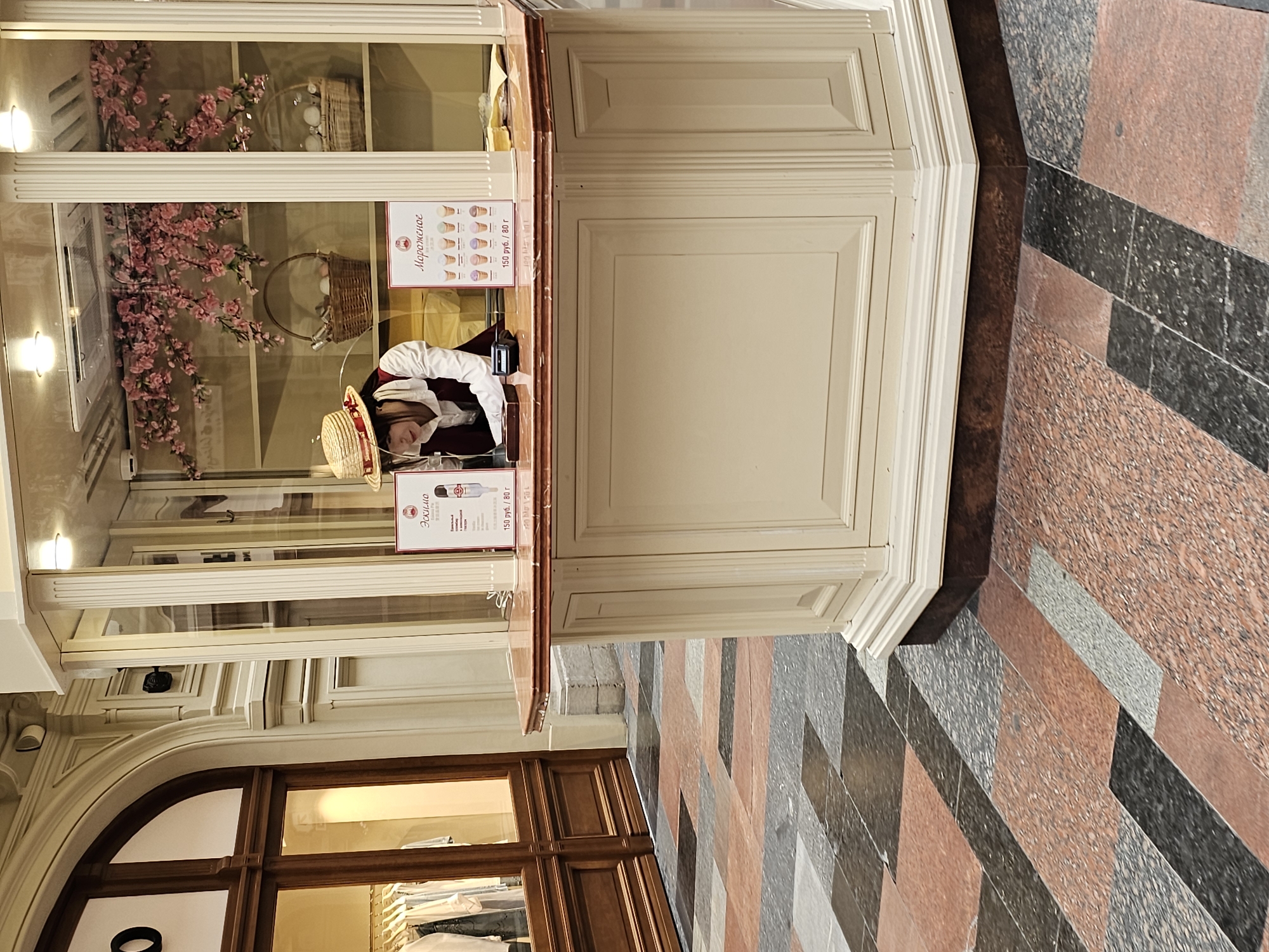 The image shows a view of a confectionery kiosk in Moscow, Russia. A female vendor wearing a straw hat and a white and maroon uniform is inside the kiosk. The kiosk has glass displays with flowers and baskets. Menu boards advertising ice cream in Russian with prices are affixed to the front of the kiosk. The kiosk is located within a large shopping area featuring decorative flooring.