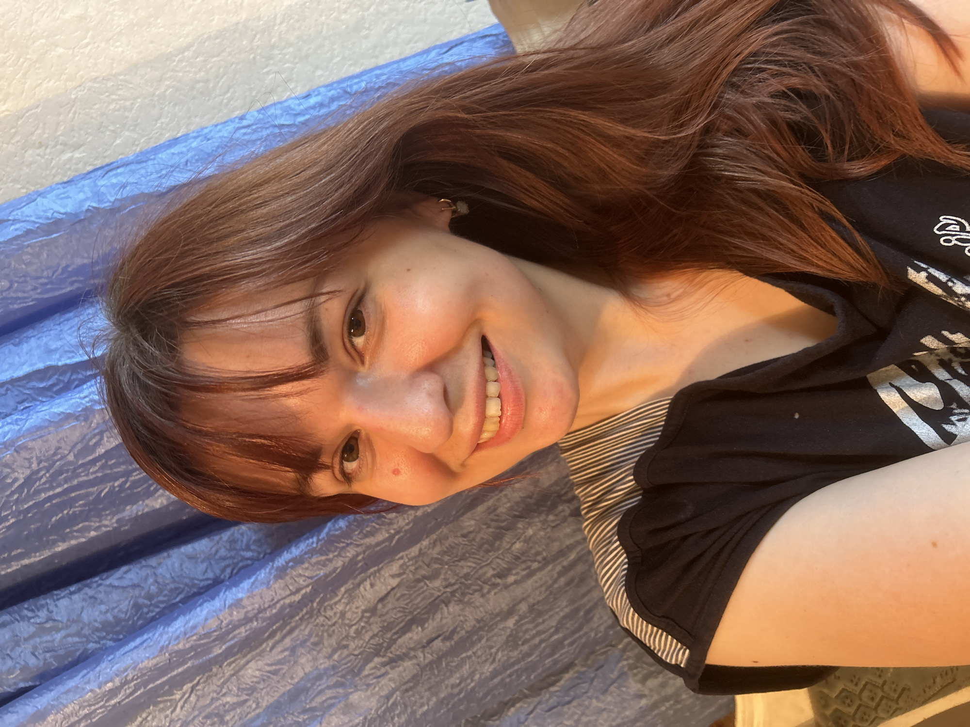 A young woman with auburn hair and bangs is smiling towards the camera. She is lying on her left side, and behind her is a wrinkled blue fabric or sheet. She is wearing a black t-shirt with a silver design on it, and a black and white striped sleeve is visible underneath the t-shirt. She is wearing a small earring in her left ear. A small mole is visible on her left cheek. The scene is indoors and well-lit.
There is no information to imply this scene is located in Фоки, Russia.