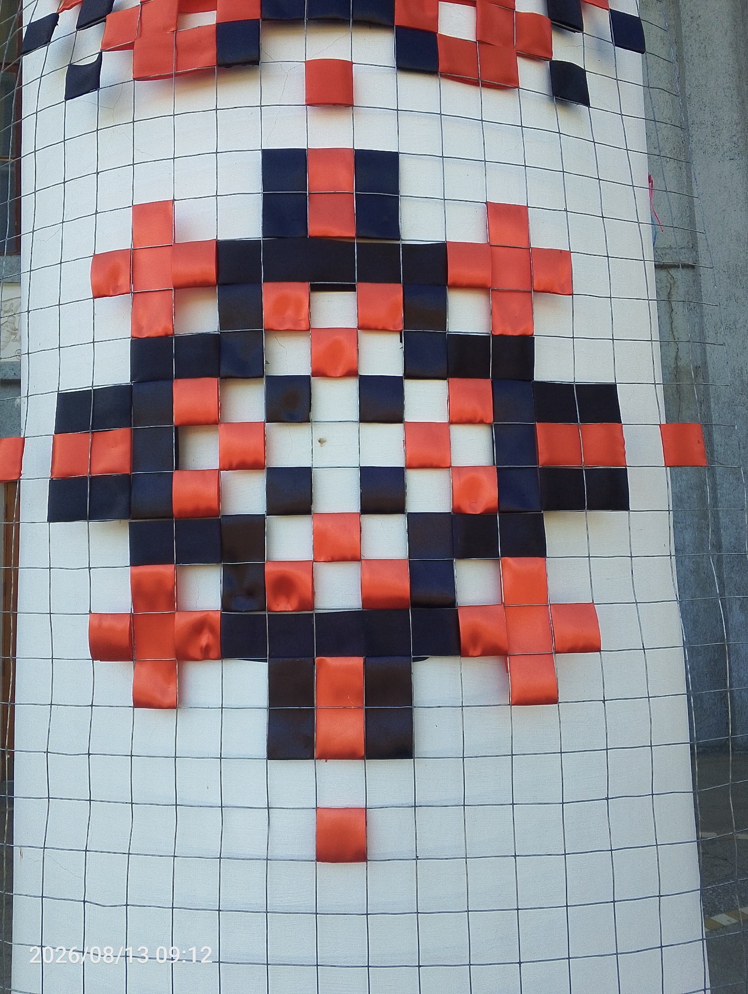 The image shows a large white cylindrical object, possibly a pillar or pole, wrapped in a grid of wire mesh. Woven through the mesh are red and black fabric squares, forming a pixelated image resembling a stylized face or character. The object is positioned outdoors, with a grey corrugated wall visible to the right and a glimpse of a building facade to the left. The ground is not clearly visible. A timestamp "2026/08/13 09:12" is present in the lower left corner. This artwork is located in Bahacheve, Ukraine.