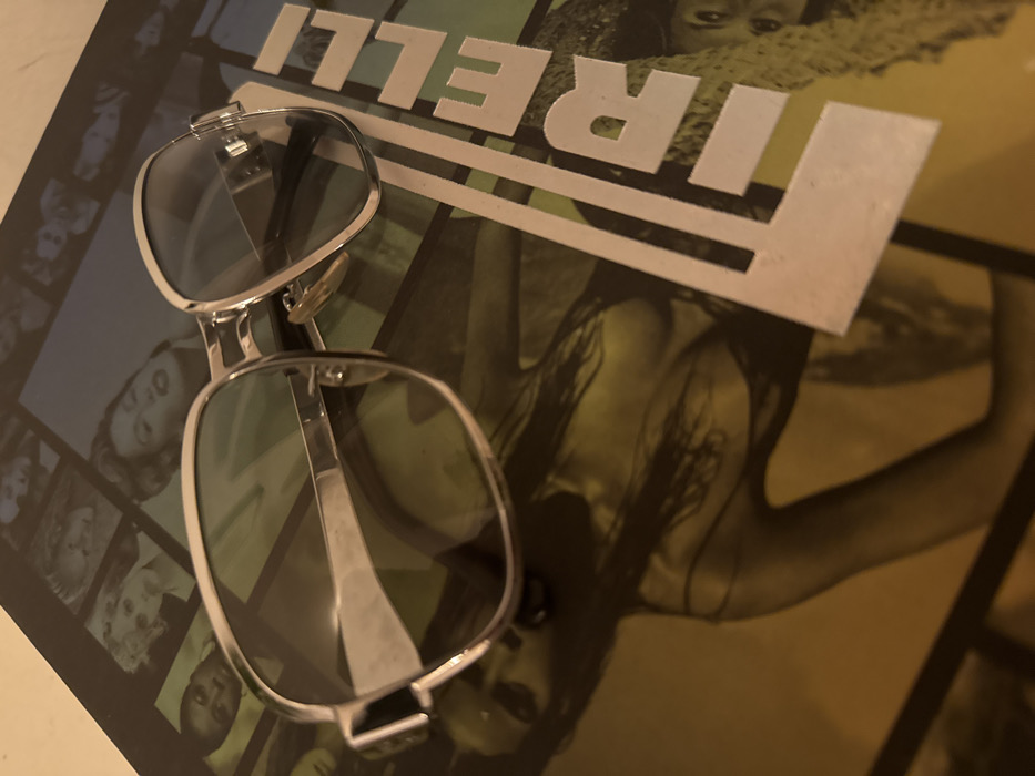 A pair of silver-toned, large-framed sunglasses with dark, possibly tinted, lenses rests diagonally on a glossy publication or poster. The publication features the word "PIRELLI" in prominent white capital letters, partially visible on the right side. The background of the publication displays a collage of photographic images, primarily featuring stylized portraits and figures of women, some in black and white, others with a yellow-green tint. One clear image shows the upper torso of a woman with long dark hair, visible beneath the left lens of the sunglasses. Reflections are visible on the metallic frames and lenses of the sunglasses. The environment is an indoor setting. There are no discernible features in the image to identify a specific location within the city of London, United Kingdom.