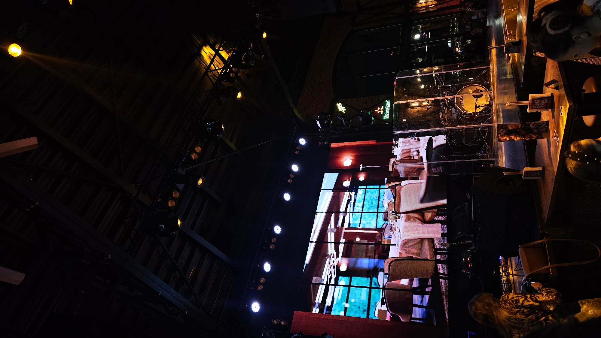 An interior shot depicts a dimly lit music venue or restaurant identified as Vintage 33 in Varna, Bulgaria. The perspective is angled upwards towards a central stage area. Overhead, an exposed metal ceiling structure supports multiple stage lighting fixtures, some emitting bright white light and others casting warm yellow beams.
The stage features a large LED screen displaying a static image of interior dining tables and chairs. In front of the screen, a drum kit, including a "Tama" branded bass drum, is positioned behind a clear plexiglass sound barrier on the right side. A green neon "Heineken" star logo is visible to the right of the screen, against a dark brick archway. Large black speakers are situated on the stage floor.
In the foreground, dark wooden tables are arranged, indicating an audience or dining section. Each table holds small white upright cardholders, likely menus or QR codes, along with other items. Two individuals are partially visible from behind in the lower frame: one on the left with long, light brown hair wearing a patterned top, looking down at the table, and another on the right showing the back of their head adorned with a decorative hair accessory. The scene suggests a live entertainment setting with a nocturnal ambiance.
