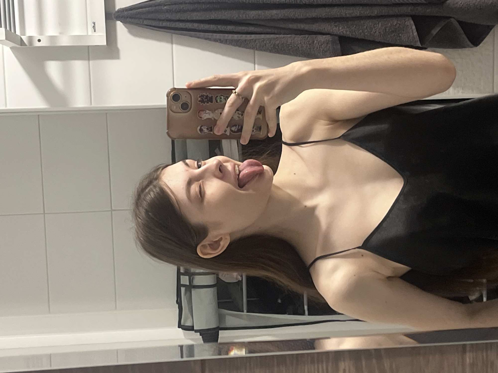 A young woman in a black tank top is taking a selfie in a bathroom. She is holding a brown iPhone with stickers on the back. Her tongue is sticking out. The bathroom has white tiles, a white cabinet, and a gray towel hanging on the wall. It's not possible to determine the location of the bathroom, or if it is in Saint Petersburg, Russia, from the provided image.
