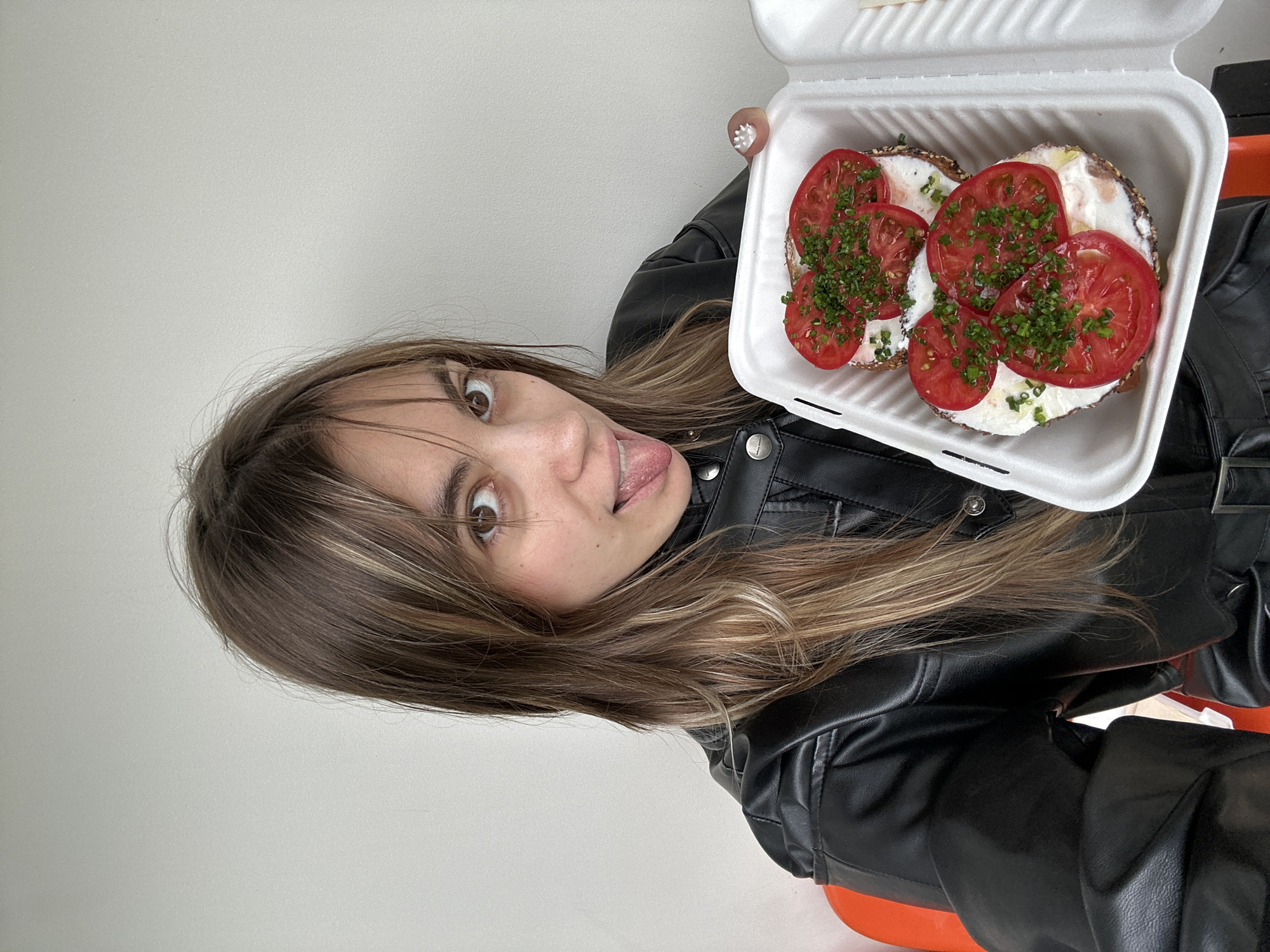 A young woman with long, blonde-streaked brown hair is pictured in a casual indoor setting, possibly a cafe or her home, in Moscow, Russia. She is wearing a black leather jacket. The woman is holding up a white takeout container filled with two bagel halves, each topped with cream cheese, sliced tomatoes, and chopped green herbs, likely chives or parsley. Her face is close to the camera, and she is making a playful expression, sticking her tongue out slightly with wide eyes. The background is a plain, light-colored wall. The lighting suggests daytime. There is no visible text in the image. The overall mood is lighthearted and informal.