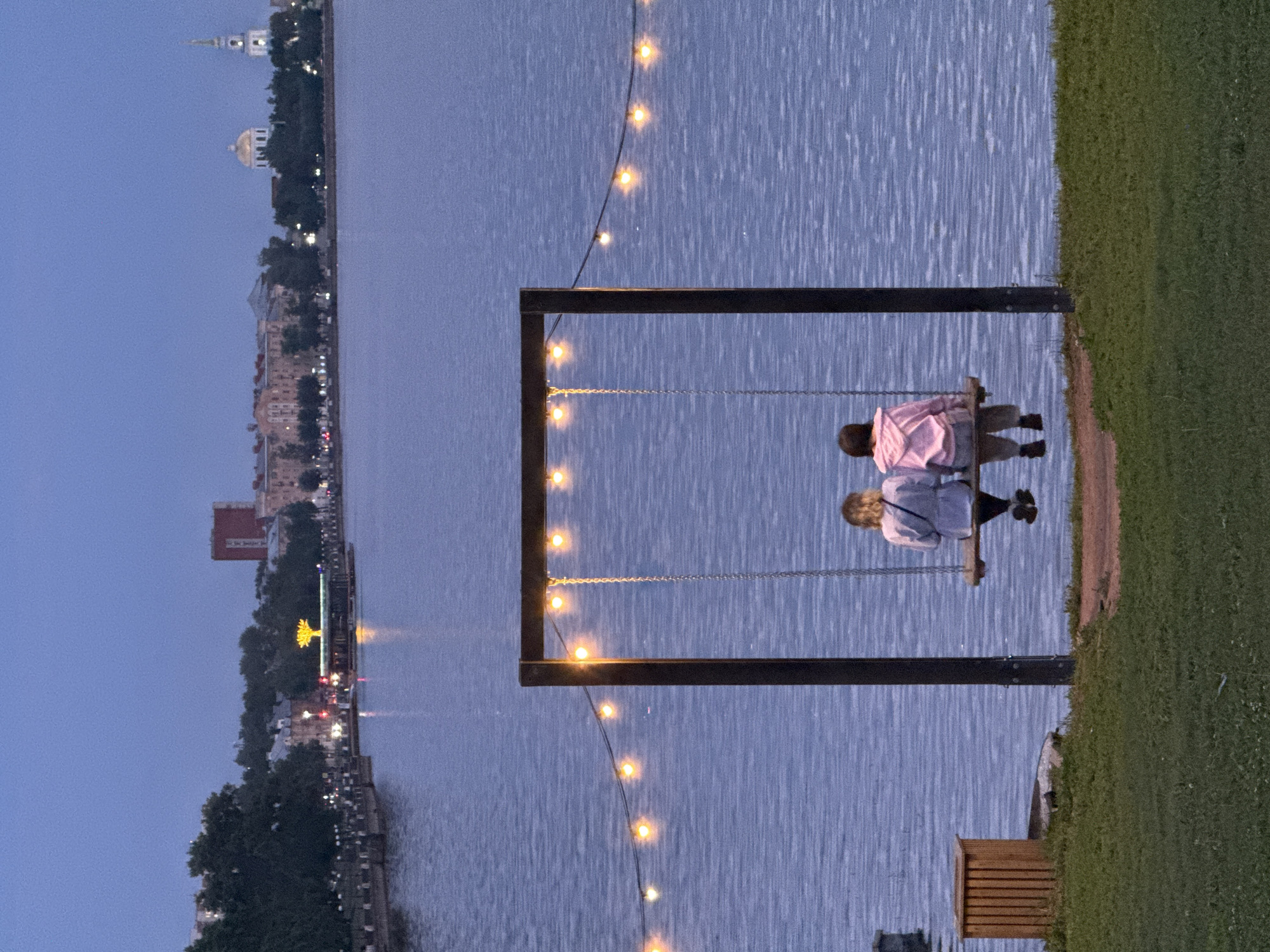 The image depicts a waterfront scene in Votkinsk, Russia, during twilight. Two individuals, seen from behind, are seated on a wooden swing suspended from a dark metal frame. They are positioned in the foreground, facing the body of water. String lights, illuminated with a warm glow, are strung across the scene above the swing and extend into the background.
The water is calm, reflecting the dusky sky and the lights. Across the water, the Votkinsk city skyline is visible. Prominent features include buildings of various architectural styles, trees lining the waterfront, and a church with a spire. A boat with illuminated decorative elements, resembling a tree, is moored near the shore. The overall atmosphere is serene and peaceful, characteristic of an evening by the water. The foreground consists of a grassy area with a wooden bin.