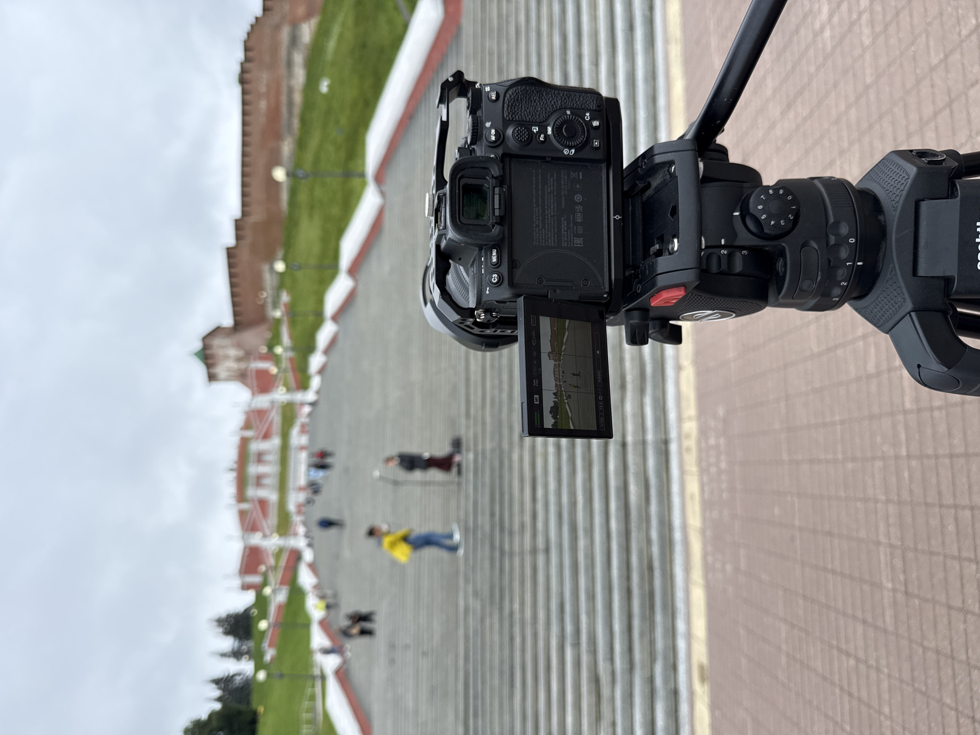 The image captures a close-up of a camera mounted on a gimbal, aimed at a wide, long staircase leading down. The camera's screen is visible, displaying a scene with people walking down the stairs. In the background, beyond the stairs, is a large, ornate red brick building, likely a historic structure, with green grass areas and lamp posts. The overall scene appears to be a public space, possibly a historical landmark or park. The weather seems overcast, indicated by the bright, diffused light and cloudy sky. Several people are scattered on the stairs, some walking, others standing, suggesting a moderately busy tourist or public area. One person in the middle ground wears a bright yellow jacket, making them stand out. The gimbal and camera suggest the media is being recorded or filmed. Text visible on the camera includes "Sony" branding and model information, as well as some production markings like "EAC" and FCC ID information. The image is oriented vertically.