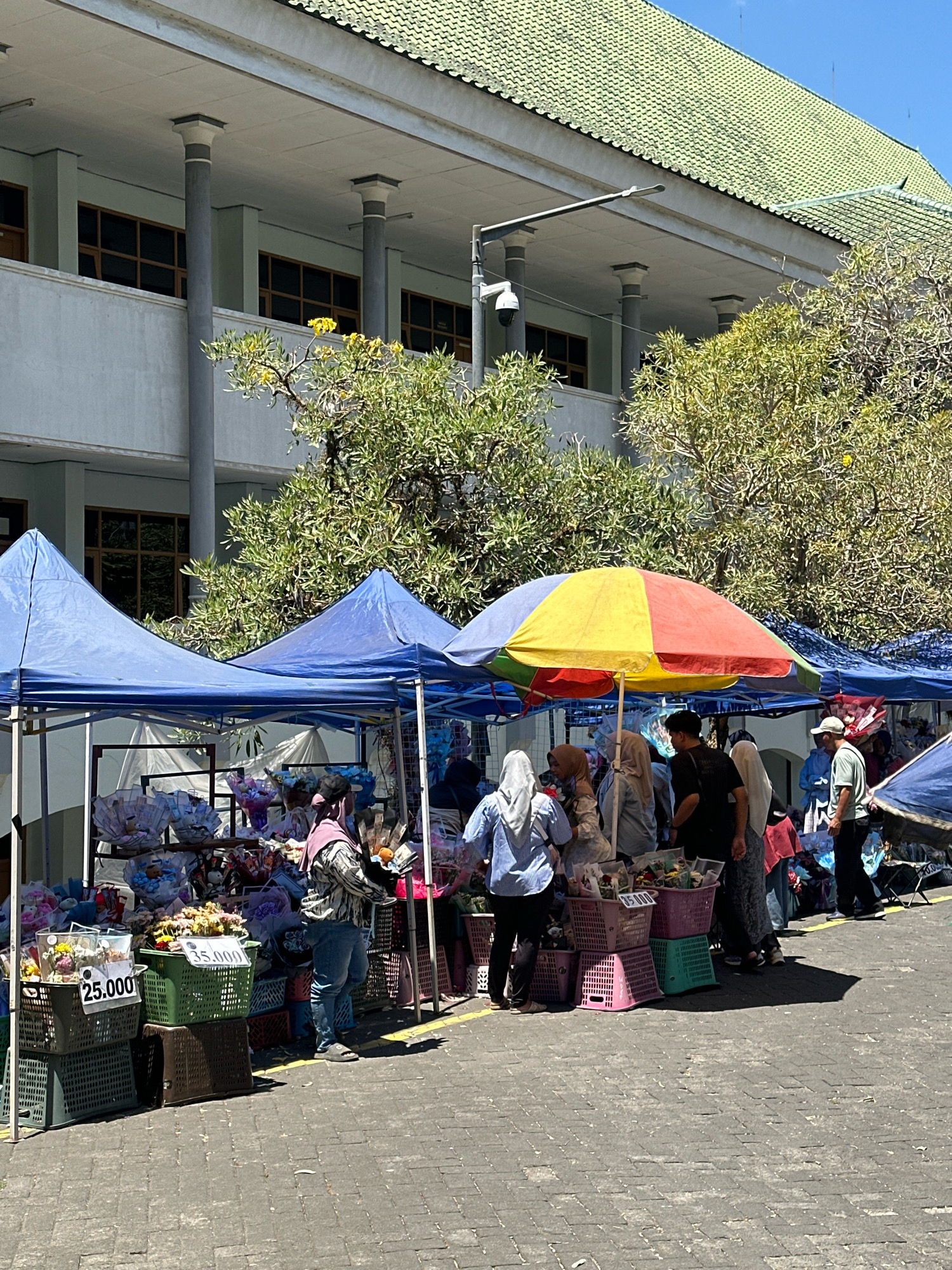A sunny outdoor marketplace is depicted in Malang, Indonesia, likely near a large building with a green tiled roof and columns. The foreground shows several market stalls covered by blue tarpaulins and a colorful umbrella. These stalls display various items, including bouquets of flowers and possibly gifts wrapped in paper and ribbons. Baskets of different sizes and colors, some containing flowers, are stacked on the ground. Price tags are visible, with "25.000" and "35.000" noted on some items. Several people, mostly women wearing hijabs, are browsing and interacting at the stalls. One person, a man wearing a light green shirt, is standing near the right side of the image. The ground is paved with interlocking stones. Trees with green foliage provide shade. A security camera is mounted on a pole in the background. The scene suggests a bustling commercial activity in a public space.