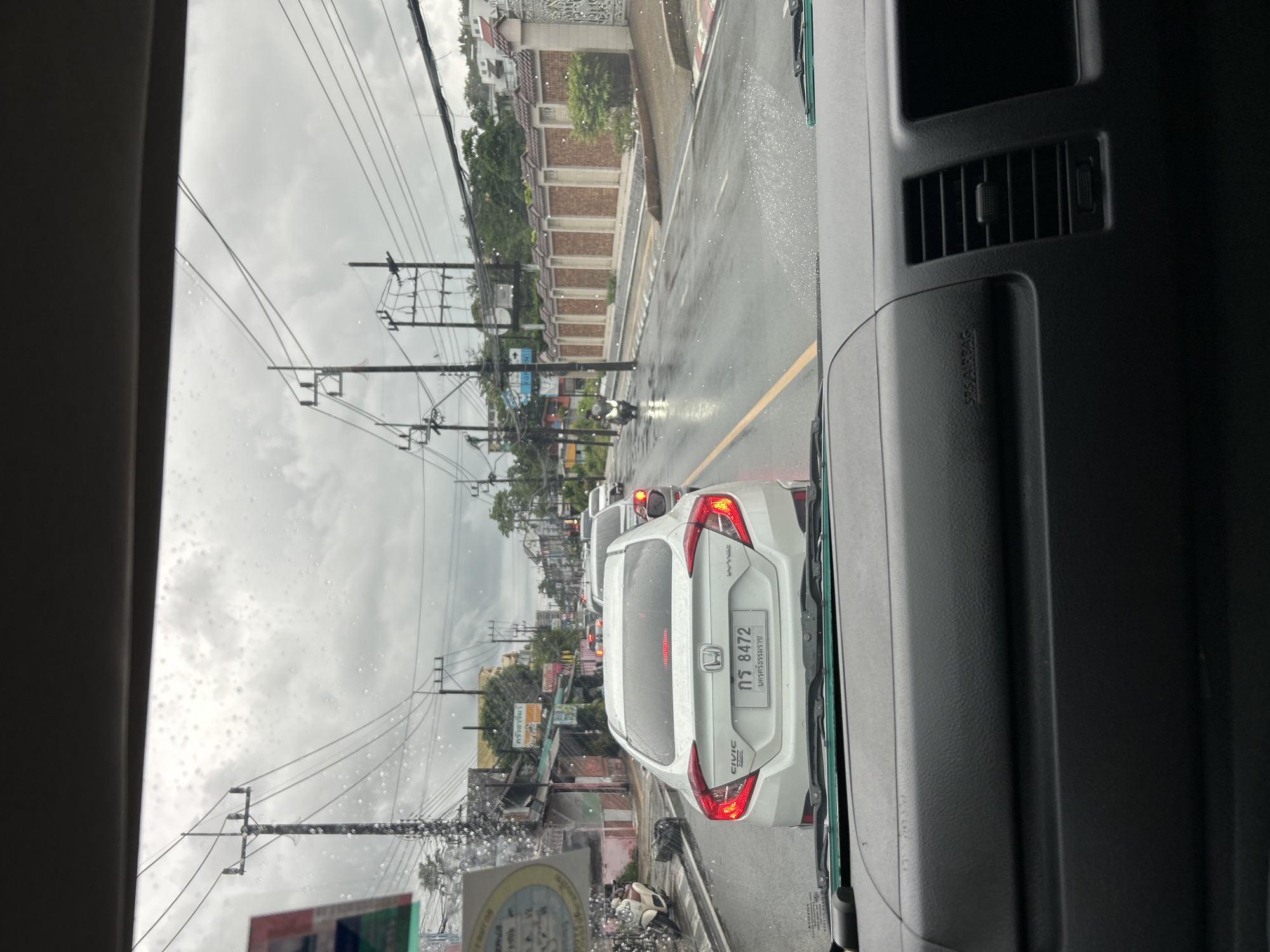 The image shows a traffic scene on a wet road, likely during or after rainfall, in Phuket City Municipality, Thailand. The perspective is from inside a vehicle, looking forward through the windshield. The road is lined with trees, buildings, and utility poles. In the foreground, a white Honda Civic is visible, with its license plate displaying "กฐ 8472". Further down the road, other vehicles, including a white van and a motorcycle, are present. The sky is overcast with gray clouds. The overall impression is one of a typical urban traffic situation in a tropical climate with rain.