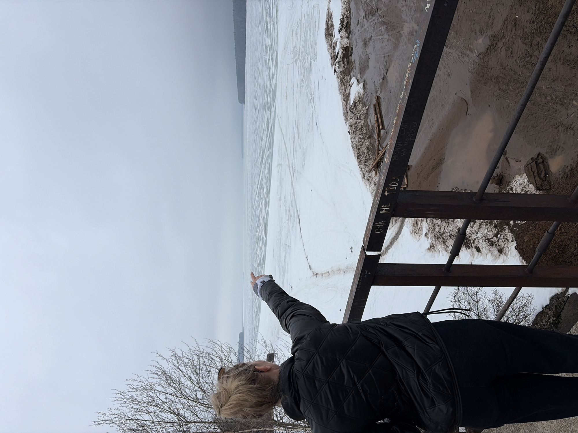 A person stands on a rusted metal bridge in Chaikovsky, Russia, pointing towards a frozen expanse of water. The horizon is obscured by a hazy, overcast sky. The ground near the bridge is a mix of snow, mud, and exposed earth. The person is wearing a black quilted jacket and dark pants. Graffiti marks the metal structure. A bare tree branch is visible near the lower left corner of the image.
