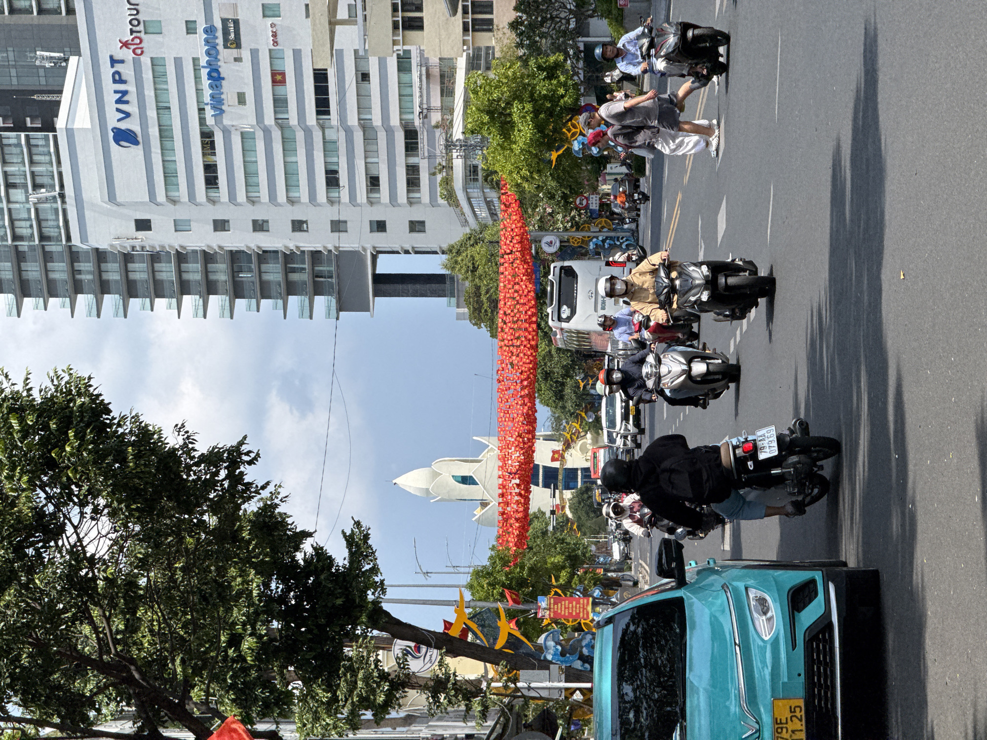 The image depicts a street scene in Nha Trang, Vietnam. A tall building labeled "VNPT" and "Vinaphone" is visible in the background. A line of red lanterns hangs above the street. Several motorbikes and a turquoise car are traveling on the road. People are walking on the sidewalk. The white, lotus-shaped Nha Trang Cathedral is also visible in the background. Trees are present on the sides of the road. The sky is light blue.