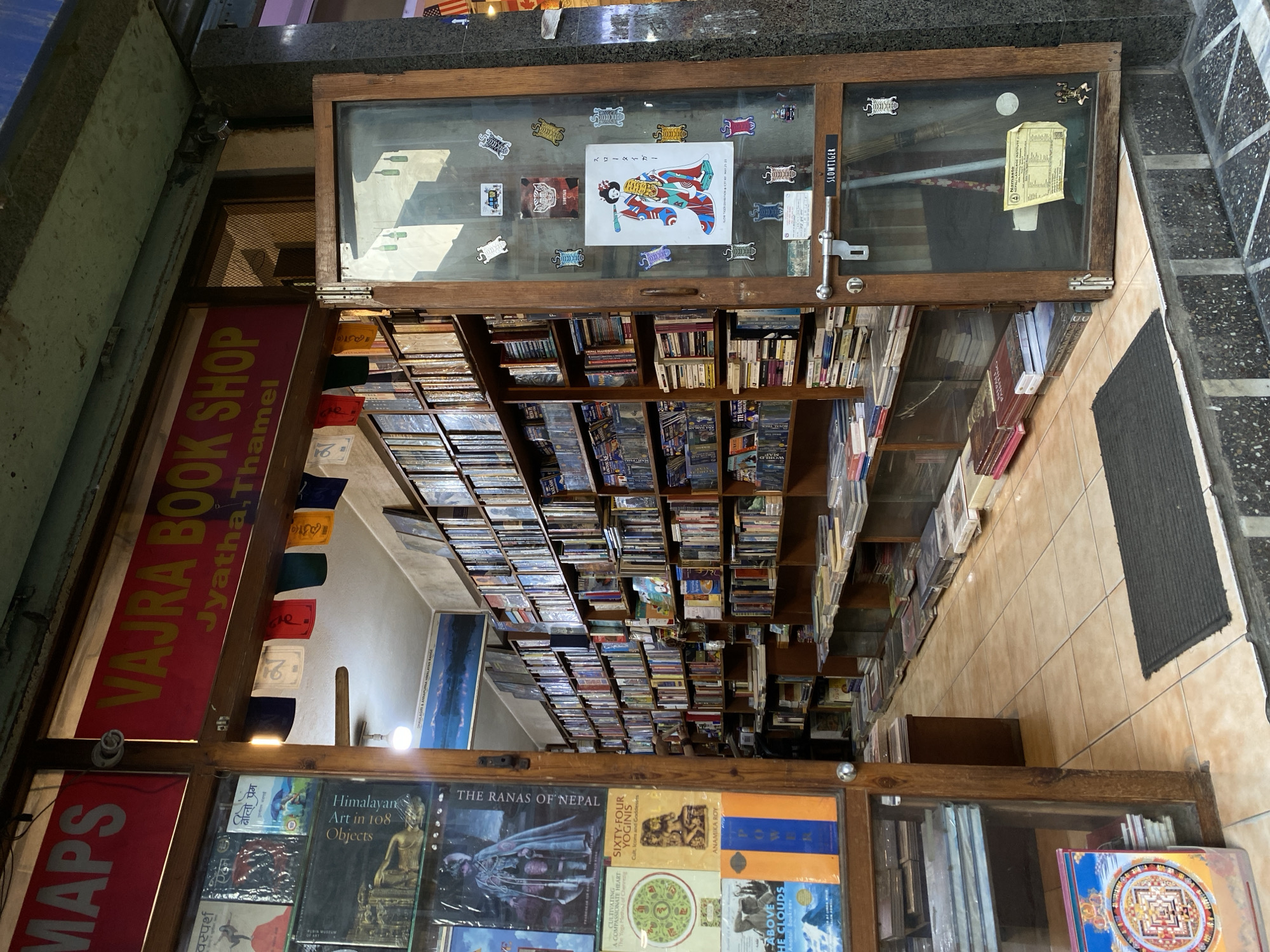 The image shows the exterior of a bookshop named "VAJRA BOOK SHOP" with "Jyatha, Thamel" written below it, located in Kathmandu Metropolitan City, Nepal. The shop entrance is open, revealing shelves packed with books. Prayer flags hang above the entrance. The shop's interior appears to be a narrow aisle lined with books on both sides. The floor is tiled. On the right side of the entrance, a glass-paneled door displays various stickers and papers. A dark doormat is placed at the entrance.