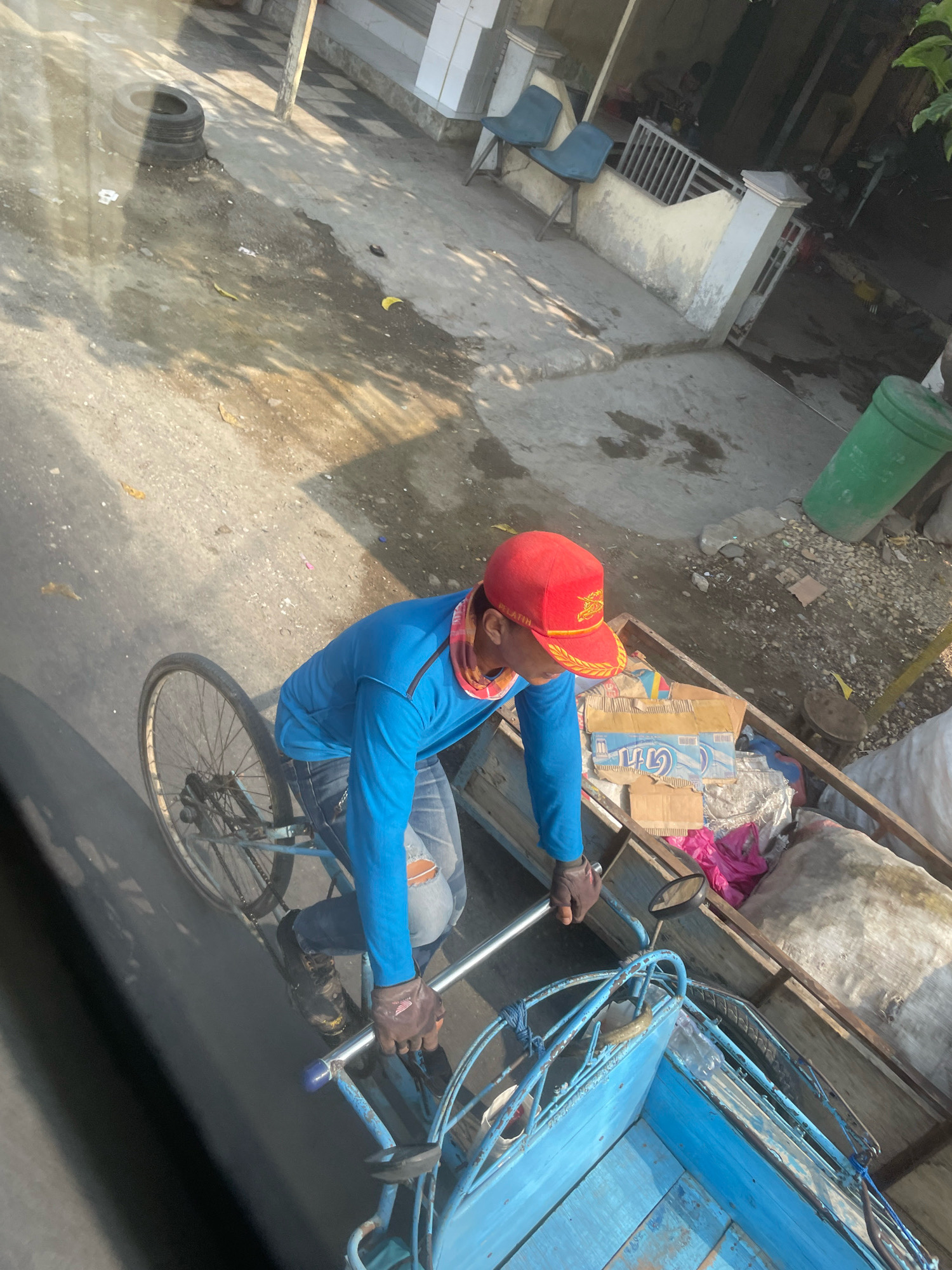 An overhead shot captures a person operating a blue tricycle laden with goods, possibly for transport, on a street in Dharma Tanjung, Indonesia. The individual, dressed in a blue long-sleeved shirt, ripped jeans, and a red cap, wears gloves and a bandana. The tricycle's cargo includes cardboard boxes, plastic bags, and sacks. The surrounding environment appears to be a street with a concrete surface, showing patches of sunlight and shadow. In the background, a green trash bin and a building with two blue chairs are visible. The scene suggests an everyday activity within a residential or commercial area.