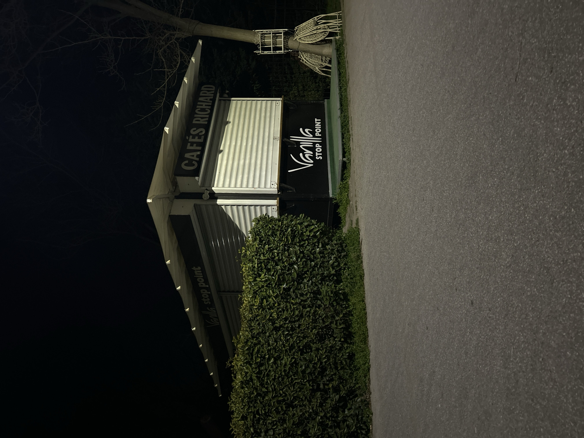 The image depicts the exterior of a small building at night, possibly a cafe or kiosk. The building has a sign that reads "CAFÉS RICHARD" and "Vanilla stop point". The shutters of the building are closed. A bush grows beside the building. Some chairs are stacked on top of each other and tied to a tree trunk. The location is possibly in the city of Varna, Bulgaria, based on the sign "Vanilla stop point". There is a paved area beside the building.