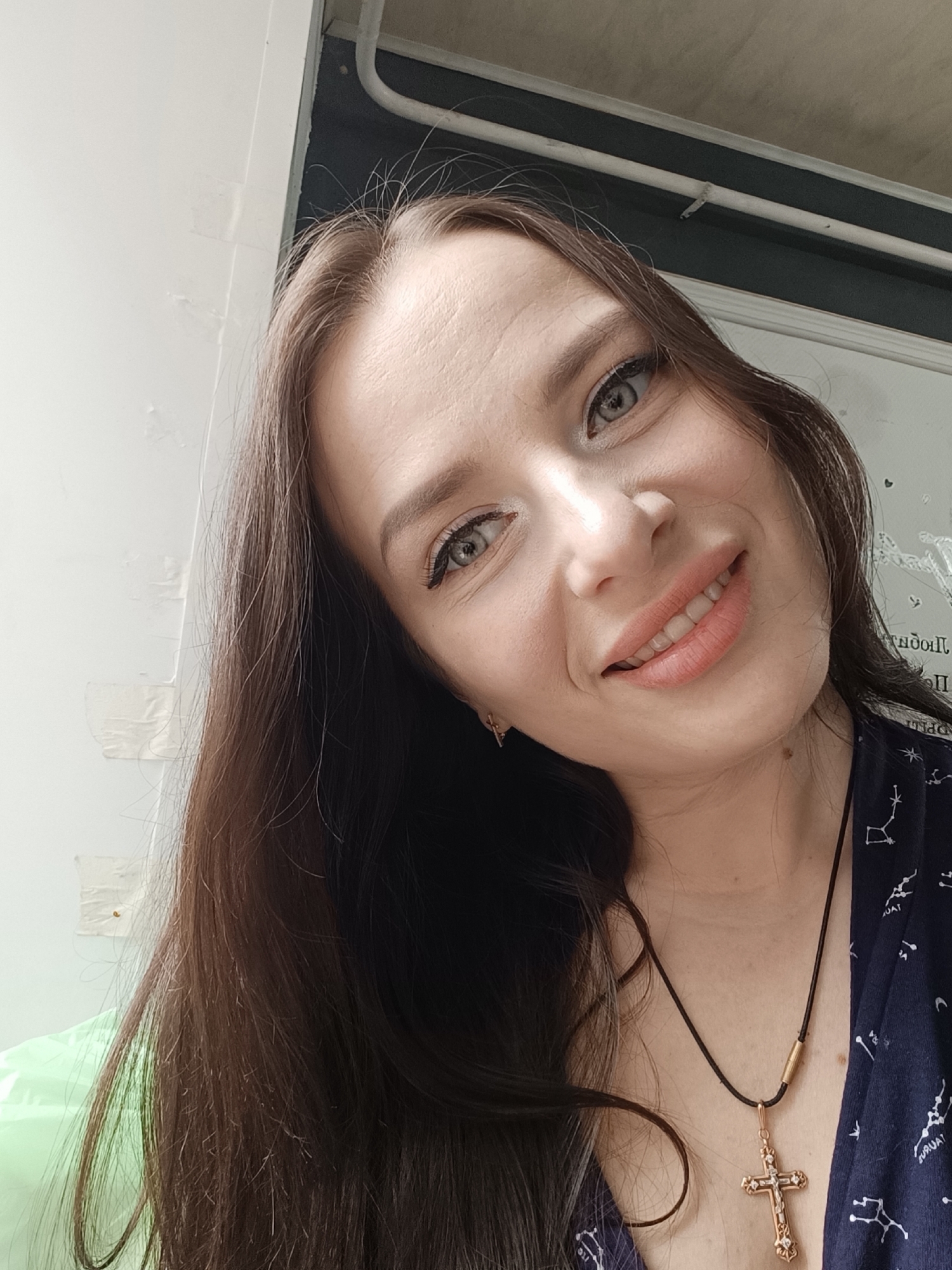 A close-up selfie depicts a fair-skinned woman with long, dark brown hair, slightly angled towards the camera. She is smiling, revealing her teeth, and her eyes are a light color. She wears what appears to be eye makeup. Around her neck, she has a black cord necklace with a gold cross pendant.
She is wearing a dark blue garment patterned with constellation designs. The background is composed of a white wall with patches of tape, and a window frame with what appears to be text on the glass, rendered backwards.
The location in the city of Rusalevka, Russia is not clear from the image.