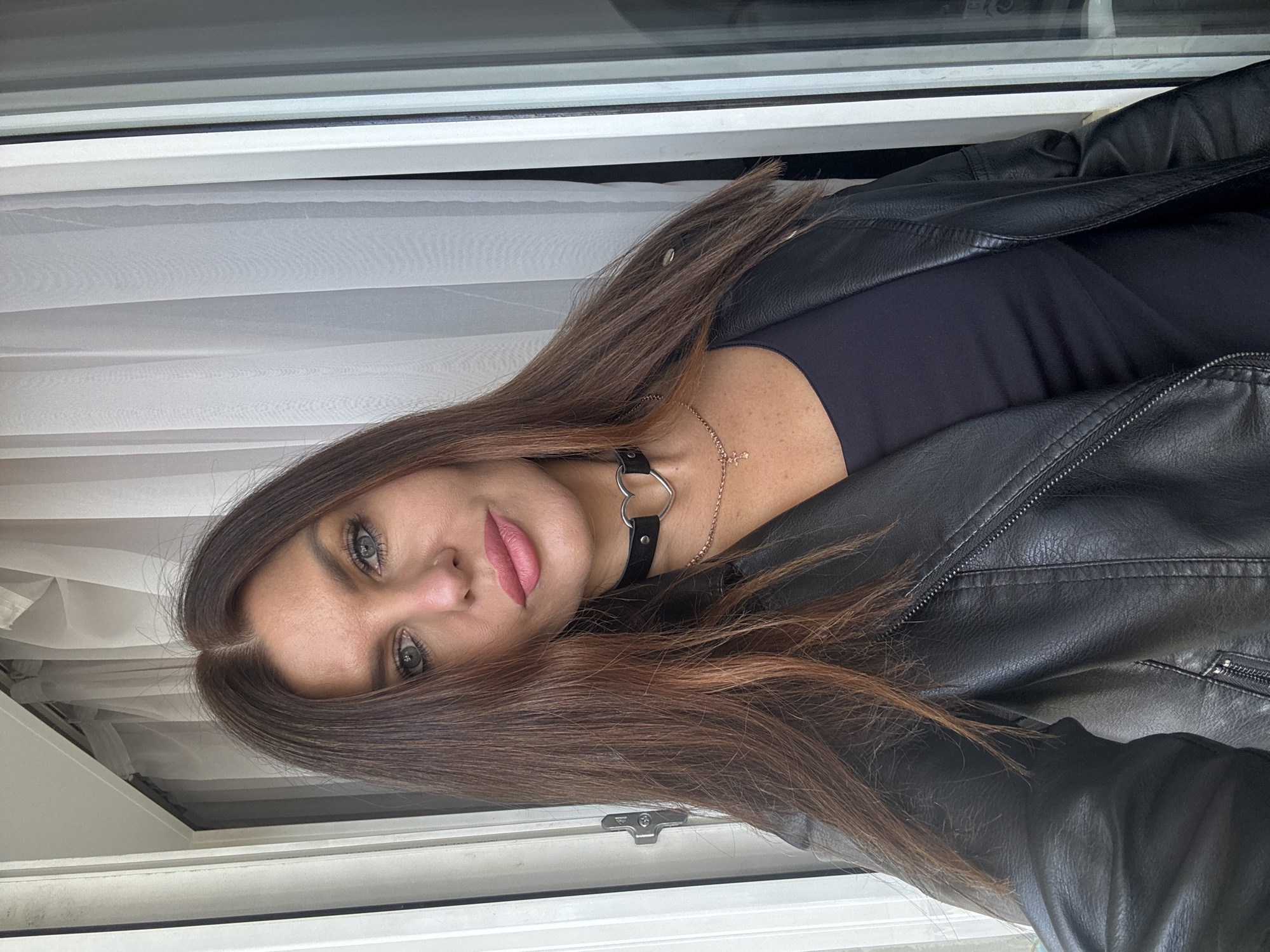 A woman with long brown hair, wearing a black leather jacket, black top, and a heart-shaped choker necklace, is positioned in front of a window with white frames and sheer curtains. The location cannot be reliably determined as Chaikovsky, Russia.