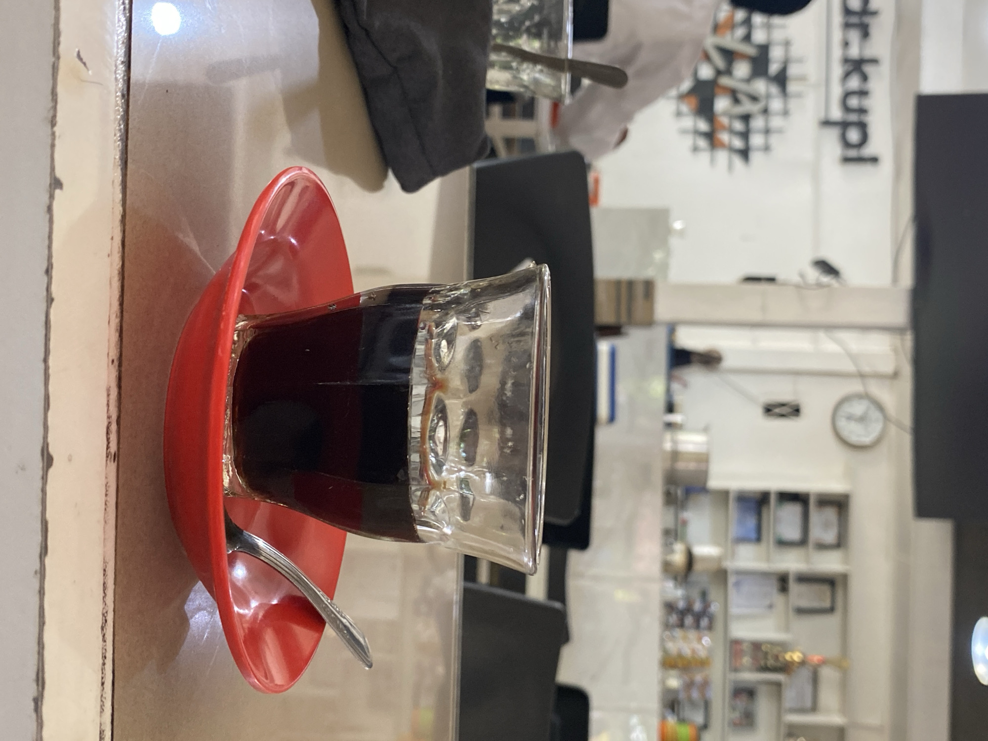 A close-up shot focuses on a clear glass of dark coffee placed on a bright red saucer on a table. A silver spoon rests on the saucer, its handle extending towards the right. In the background, out of focus, a person in a white shirt is visible, as are tables and chairs, suggesting an indoor setting such as a cafe. The cafe appears to be in Lhokseumawe, Indonesia, as indicated by the text "dr. kupl" on the wall behind the person. The overall lighting suggests daytime.