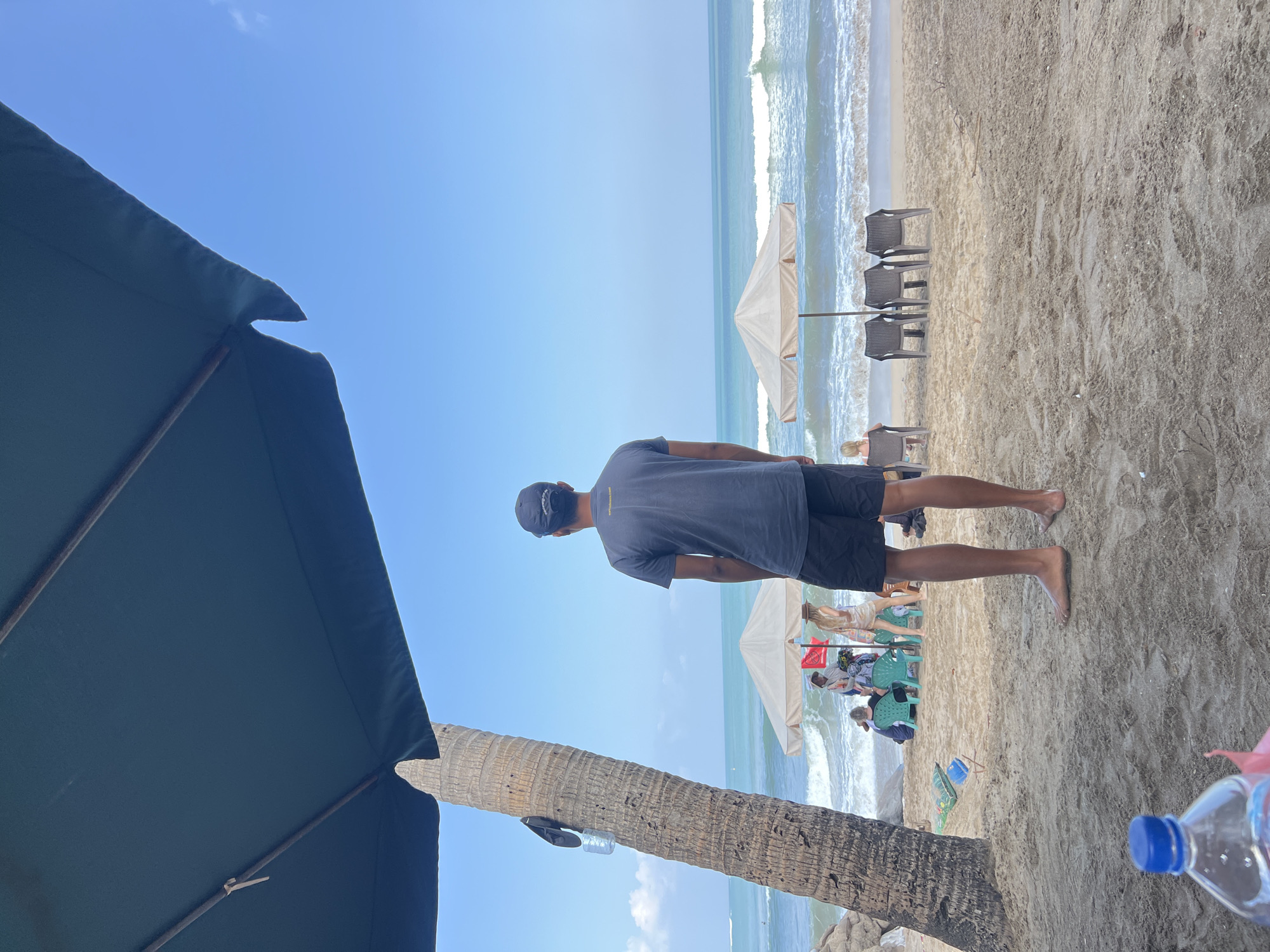 A man stands on a sandy beach in Kuta, Indonesia, facing the ocean. He is wearing a dark grey t-shirt and dark shorts, and a baseball cap. The man is positioned under a large, dark green umbrella, with the trunk of a palm tree to his left. In the background, the turquoise ocean stretches to the horizon under a clear blue sky. Gentle waves are breaking on the shore. Further back on the beach, two cream-colored umbrellas are set up, providing shade for several people sitting on chairs. A few lounge chairs are also visible near the water. The sand appears light brown and textured, with some scattered debris. In the foreground, the edge of another large dark green umbrella is visible at the top of the frame, and a partially visible blue-capped water bottle rests on the sand.