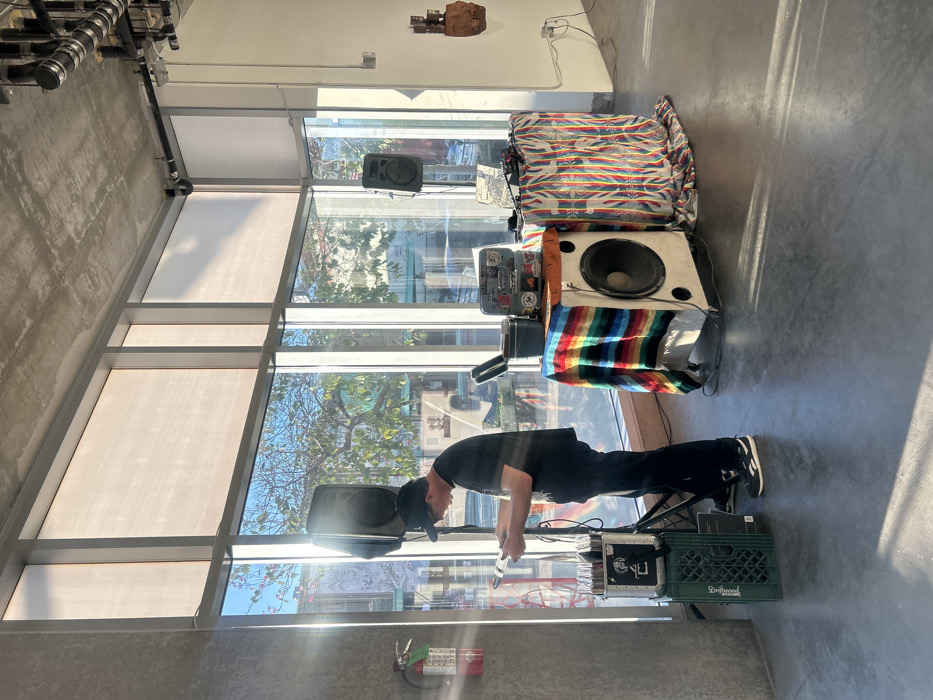 An adult male stands within a modern, urban interior in Los Angeles, United States of America. The room features floor-to-ceiling windows on two walls, revealing an urban street scene with trees, buildings, and parked cars outside. Sunlight streams through the windows, illuminating a polished concrete floor and an exposed concrete ceiling with industrial piping.
The male, dressed in a black t-shirt, black pants, black baseball cap worn backward, and black-and-white sneakers, is positioned on the left, examining vinyl records or sleeves held in his hands. To his right is a DJ setup. This setup consists of a mobile table draped with a multi-colored striped blanket, supporting a large white speaker cabinet, an open, sticker-covered metal case, and audio mixing equipment with visible cables. Two black PA speakers on stands flank the setup; one is to the male's immediate left, and the other further right. A green plastic milk crate filled with records sits on the floor below the left PA speaker stand. A red fire extinguisher is mounted on a concrete pillar to the far left. On the right wall, a sculptural object made of dark wood and metal is affixed.