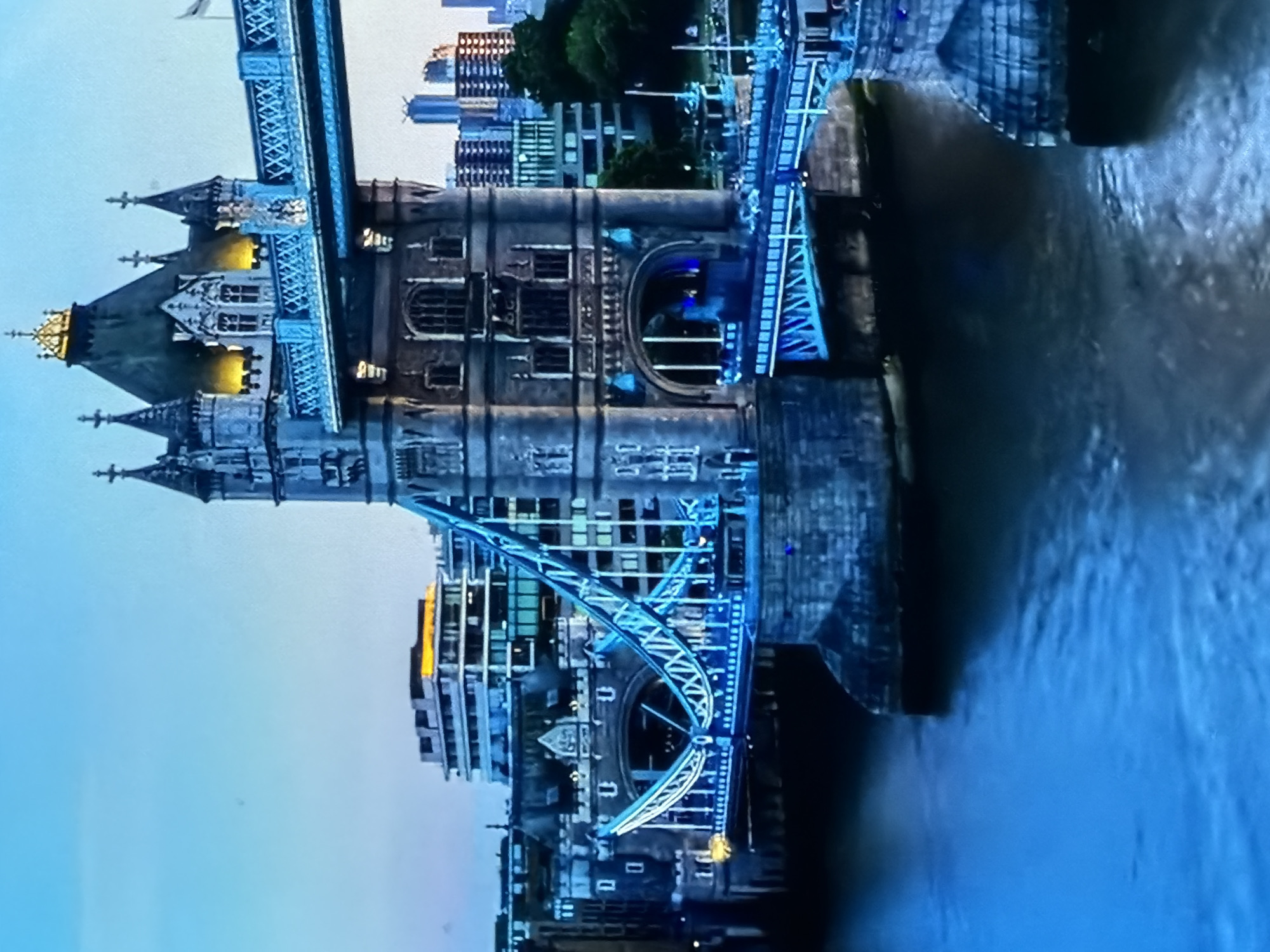 The image presents an elevated, detailed view of Tower Bridge, illuminated against a twilight sky. The two principal towers, constructed in Victorian Gothic style, feature prominent warm golden lighting on their upper sections and rooftops, contrasting with the vibrant blue illumination outlining their lower structures, arches, and the pedestrian walkways connecting them. The bridge's road decks are also highlighted with blue lights, showcasing the intricate truss work of the bascules, particularly on the left side where the deck appears to curve upwards. The dark, flowing water of the River Thames occupies the foreground and flows beneath the bridge. In the background, various urban buildings are visible, some with illuminated windows, contributing to a bustling cityscape backdrop. No specific individuals or active movements are discernible within the frame. This scene is located in the city of Maidstone, United Kingdom.