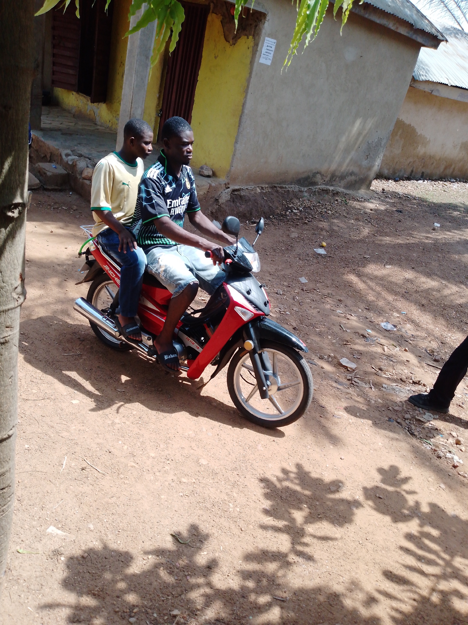 The image shows two young men on a red and black motorbike, parked on a dirt road. The rider is wearing a dark sports shirt and shorts, while the passenger is wearing a light-colored shirt and jeans. The background includes a yellow building with a red door and another building with a corrugated roof. The location is unidentified.