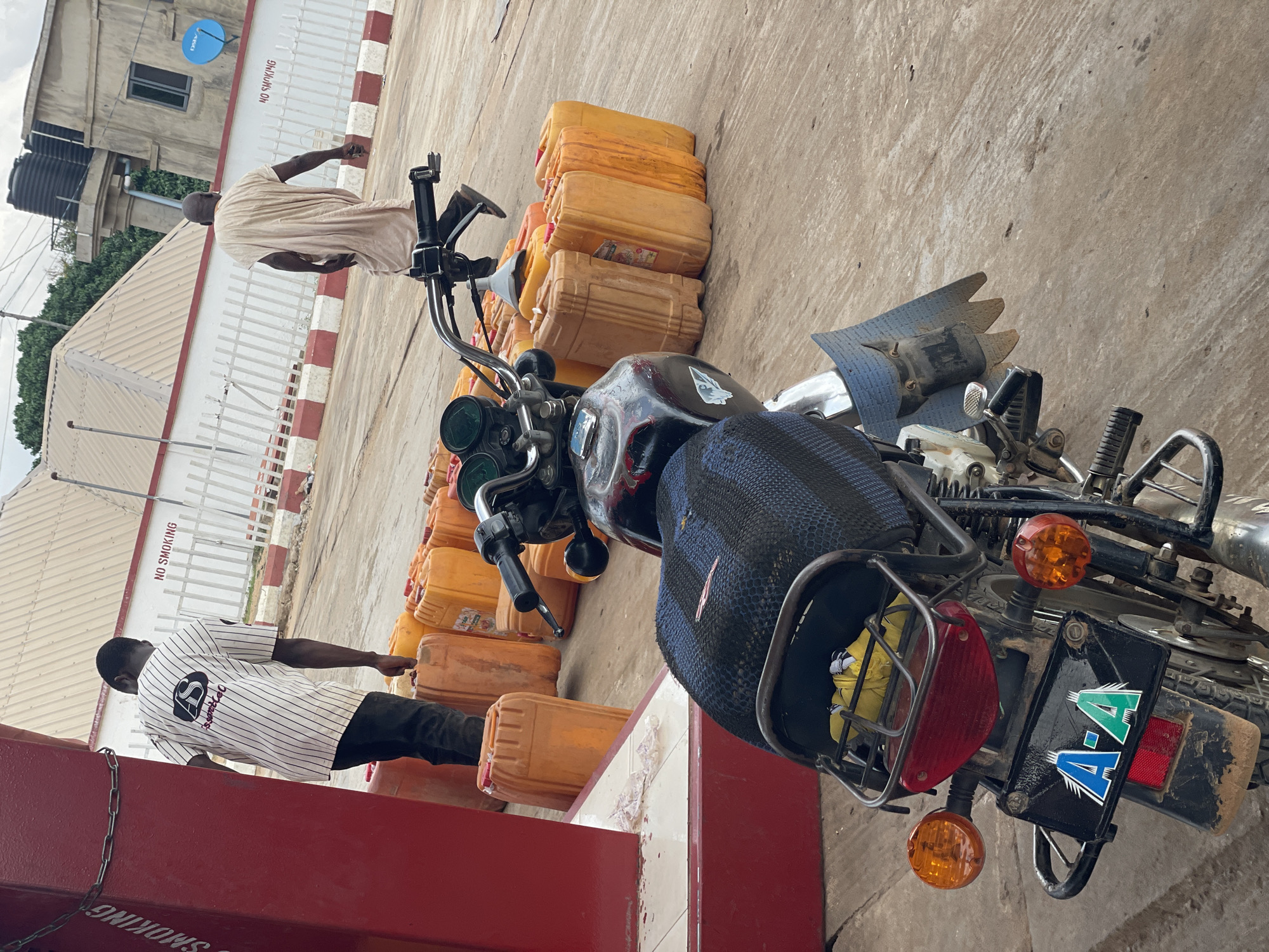 The image depicts a motorcycle heavily laden with multiple yellow jerrycans, presumably containing fuel, at what appears to be a petrol station. Two men are present: one dressed in a light-colored traditional garment standing behind the motorcycle, and another in a striped shirt and dark trousers near the front of the bike. The motorcycle has a "A-A" license plate. The petrol station background includes a building with a water tank and "NO SMOKING" signs on the fence. The location is presumably Akwanga, Nigeria. The scene suggests informal fuel sales or transport.