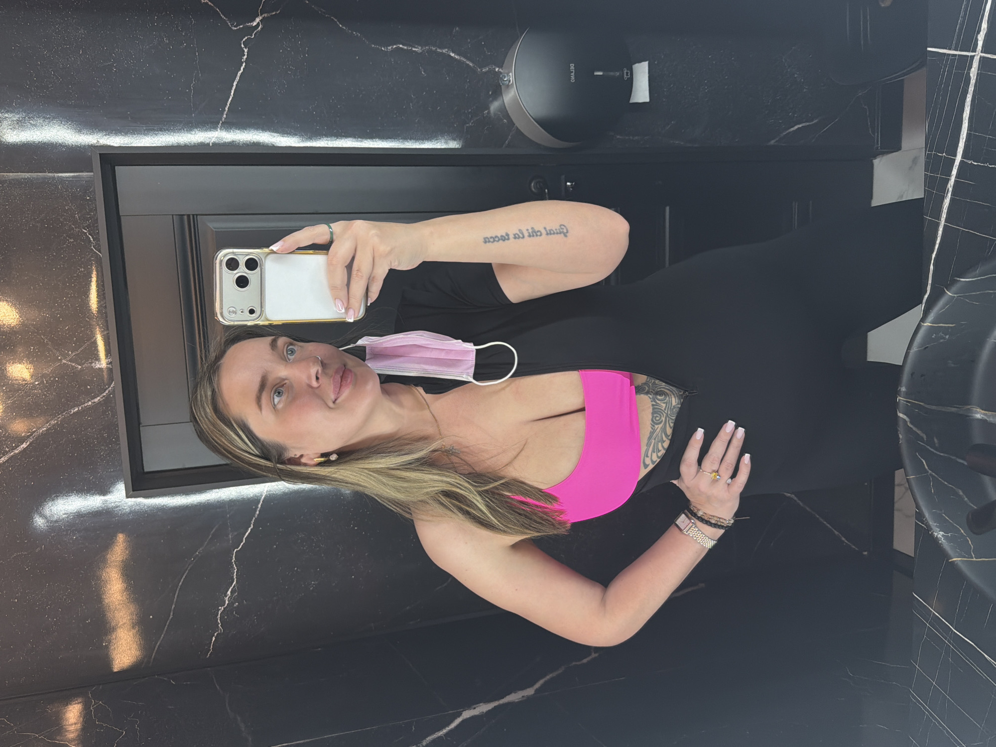 A woman is taking a selfie in what appears to be a bathroom with black marble walls and a black door. She is wearing black leggings, a black top, and a bright pink sports bra. She has blonde hair and fair skin. In her left hand, she holds a smartphone with a clear case. A pink disposable face mask hangs loosely around her neck. The background includes a soap dispenser on the wall and a sink basin to the right. The image is angled, suggesting it was taken in a mirror. No specific details indicate that it is in Moscow, Russia.