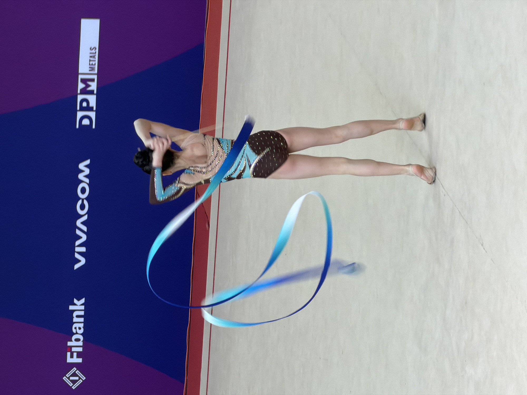 This media file depicts a female rhythmic gymnast performing a routine with a ribbon in an indoor sports arena. The gymnast, positioned towards the center-right of the frame, wears a dark leotard with intricate blue and teal patterns and sparkling embellishments. Her arms are raised, actively maneuvering a long, dual-toned ribbon that transitions from dark blue to light blue, creating dynamic, flowing loops and trails due to its motion. Her legs are extended gracefully, and she wears light-colored rhythmic gymnastics slippers.
The performance takes place on a light-colored mat or floor. On the left side of the image, a large advertising banner or wall is visible, predominantly dark blue with a subtle purple gradient. This banner features several clearly legible sponsor names: "Fibank" (with its logo), "VIVACOM", and "DPM METALS" (with "METALS" oriented vertically next to "DPM"). The overall setting suggests a competitive event or demonstration in Sofia, Bulgaria, as indicated by the context. The lighting is bright and even, typical of an indoor sports venue. The gymnast's pose conveys grace and concentration, central to the art of rhythmic gymnastics.