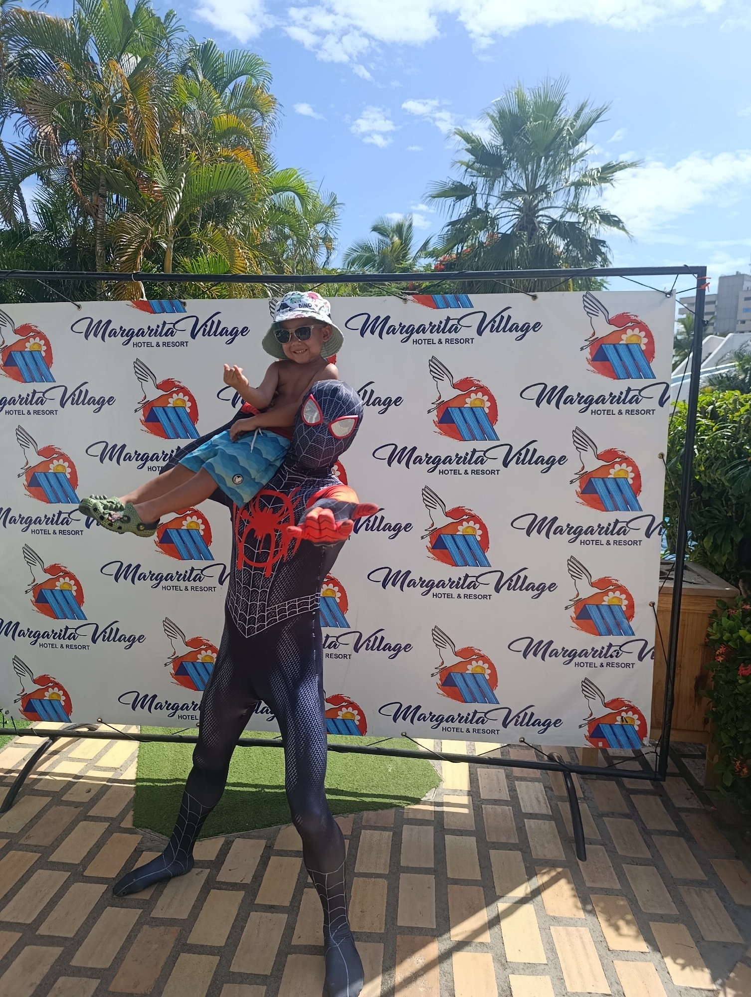 Here's a detailed description of the image:
**General Scene:**
The image is taken outdoors during the day. The background features lush tropical foliage, palm trees, a clear blue sky with scattered clouds, and a building in the distance.
**Subjects:**
* A person dressed in a Spiderman costume is holding a young child on their shoulders. The Spiderman costume appears to be a version with black and red details.
* The child is wearing a hat, sunglasses, and swim trunks. They are giving the thumbs-up sign with one hand.
**Objects:**
* A backdrop or banner stands behind the subjects. It prominently features the "Margarita Village HOTEL & RESORT" logo, repeated several times. The logo includes a graphic with a bird and a sun/flower image.
* The ground is paved with brown tiles and green artificial turf in one small area.
**Actions/Interactions:**
* Spiderman is holding the child on their shoulders, suggesting a playful or posed interaction.
* The child is smiling and giving the thumbs-up, indicating enjoyment.
**Environment:**
The scene seems to be set in a resort or tourist area, indicated by the "Margarita Village HOTEL & RESORT" banner and the tropical landscaping.
**Location:**
The location is implied by the "Margarita Village" name. Margarita Island is in Venezuela.
It's not possible to pinpoint an exact address or neighborhood in the city of Porlamar without additional information.