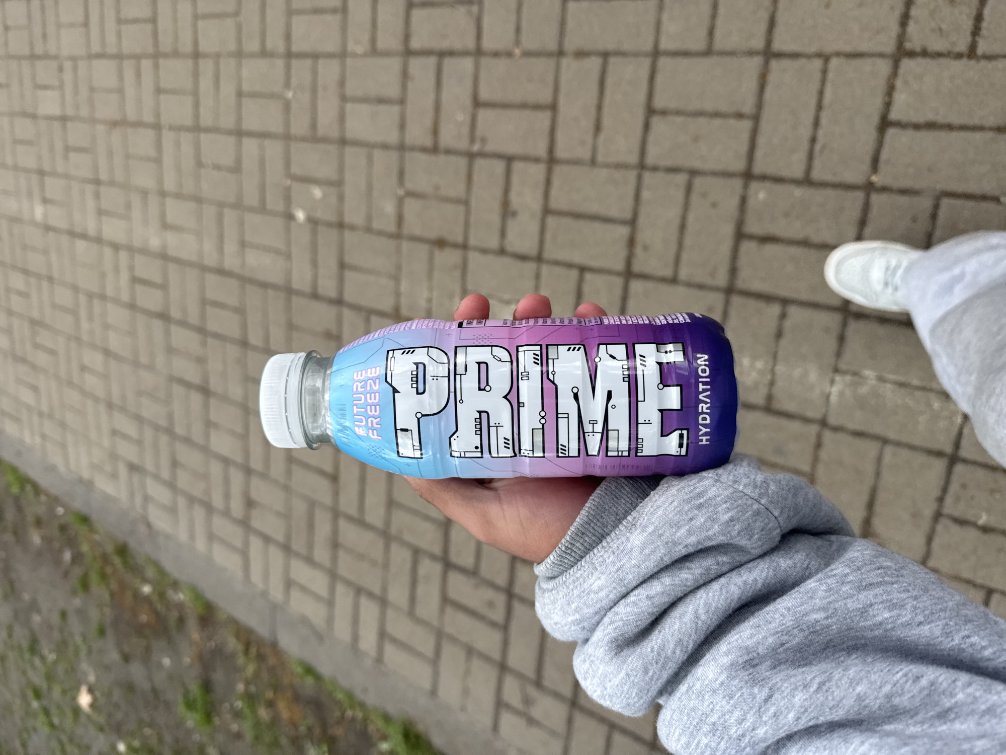 The image captures a person's right hand holding a "PRIME HYDRATION" bottle outdoors in Yerevan, Armenia. The hand is clad in the light grey sleeve of a hooded sweatshirt. The bottle, featuring a white cap, displays a colorful label with a blue-to-purple gradient. The prominent brand name "PRIME" is written in a futuristic, circuit-board-like font across the center of the label, with "HYDRATION" visible beneath it. Vertically oriented on the blue section of the label, the flavor "FUTURE FREEZE" is clearly legible.
The setting is a paved outdoor area, likely a sidewalk or pedestrian path, constructed from light-colored, rectangular interlocking blocks. A patch of green vegetation, possibly grass or weeds, is visible along the left edge of the pavement. In the background to the right, a person's lower leg and foot, wearing a white sneaker and grey pants, are partially visible, indicating they are either standing or walking. The scene is illuminated by natural light, suggesting it is daytime with an overcast or diffused sky, as no strong shadows are apparent. No specific emotions are discernible from the partial view of the person.