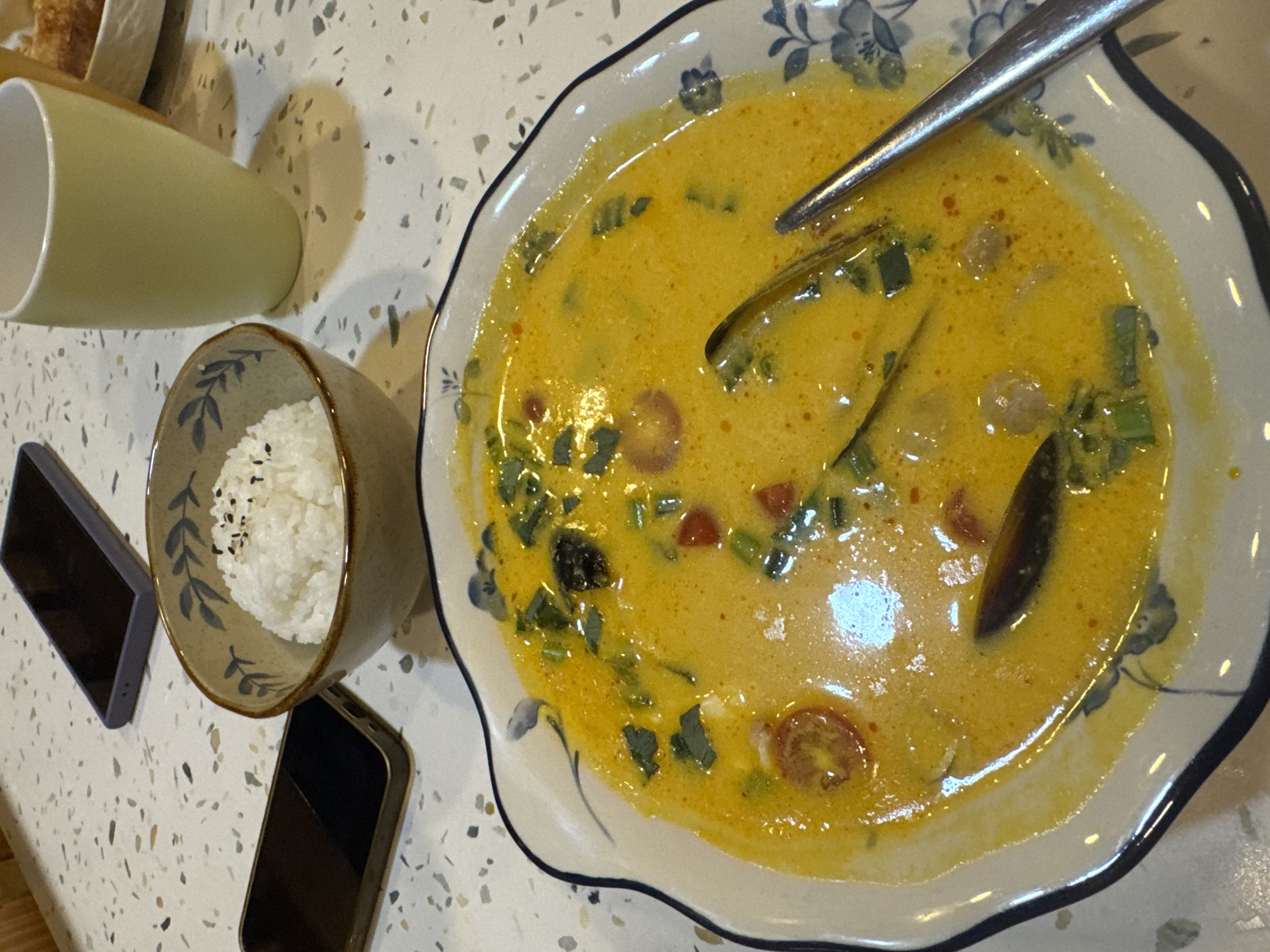 The image shows a top-down view of a meal laid out on a speckled countertop, likely in a restaurant or home setting in Bac Nha Trang Ward, Vietnam. The primary subject is a bowl of vibrant yellow curry soup, featuring visible pieces of green onions, cherry tomatoes, and what appear to be mussels or other seafood. A silver spoon rests in the soup. To the left of the soup, a small bowl of white rice is visible, topped with black sesame seeds. A pale green ceramic cup is also present on the left side of the frame. Two dark-colored smartphones are positioned below the rice bowl, one partially obscured by the other. The overall impression is of a casual meal being enjoyed. The lighting suggests it could be daytime, but there are no clear indicators of the weather or specific time. The setting is clean and well-lit. There is no visible text in the image.