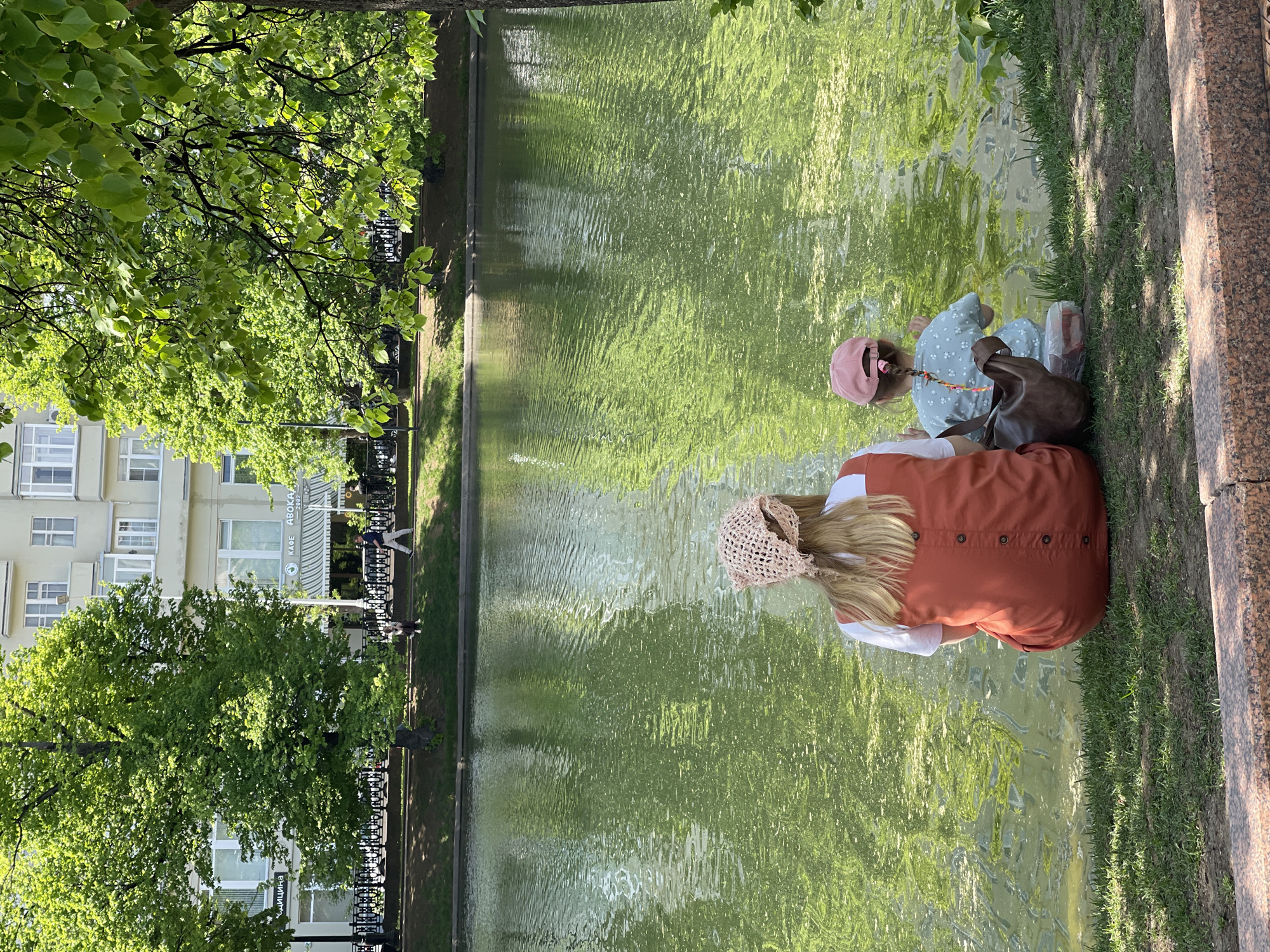 Two females are at the edge of a pond in the Unknown city of Unknown. One is wearing an orange dress with a cream colored hat. The other is wearing a dress with white flowers and a pink hat. A dark brown bag is next to the second female. Behind the pond is a building with a restaurant on the bottom floor. A black fence is between the pond and the building.