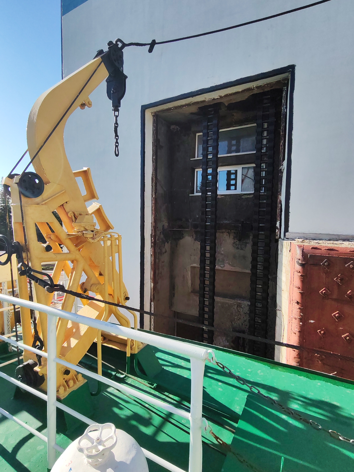 The image shows a scene on what appears to be a vessel, likely a ship or large boat, with a yellow crane apparatus prominently positioned in the foreground. The deck is green, with a white railing along the side. A chain is loosely draped across the deck.
Behind the crane, a large, rectangular opening or doorway is visible in the side of a building. The interior of the opening appears dark and contains at least one window, reflecting a building across the way. The doorway is flanked by two vertical black, track-like components on the inside. To the right of the doorway is a rusted red section of the building's exterior. The primary building surface is white or light grey.
The location is identified as Белоусово, Russia, though the specific context within the city is not apparent. There is no evidence of human presence directly in the image, though the vessel implies a maritime or waterside context.