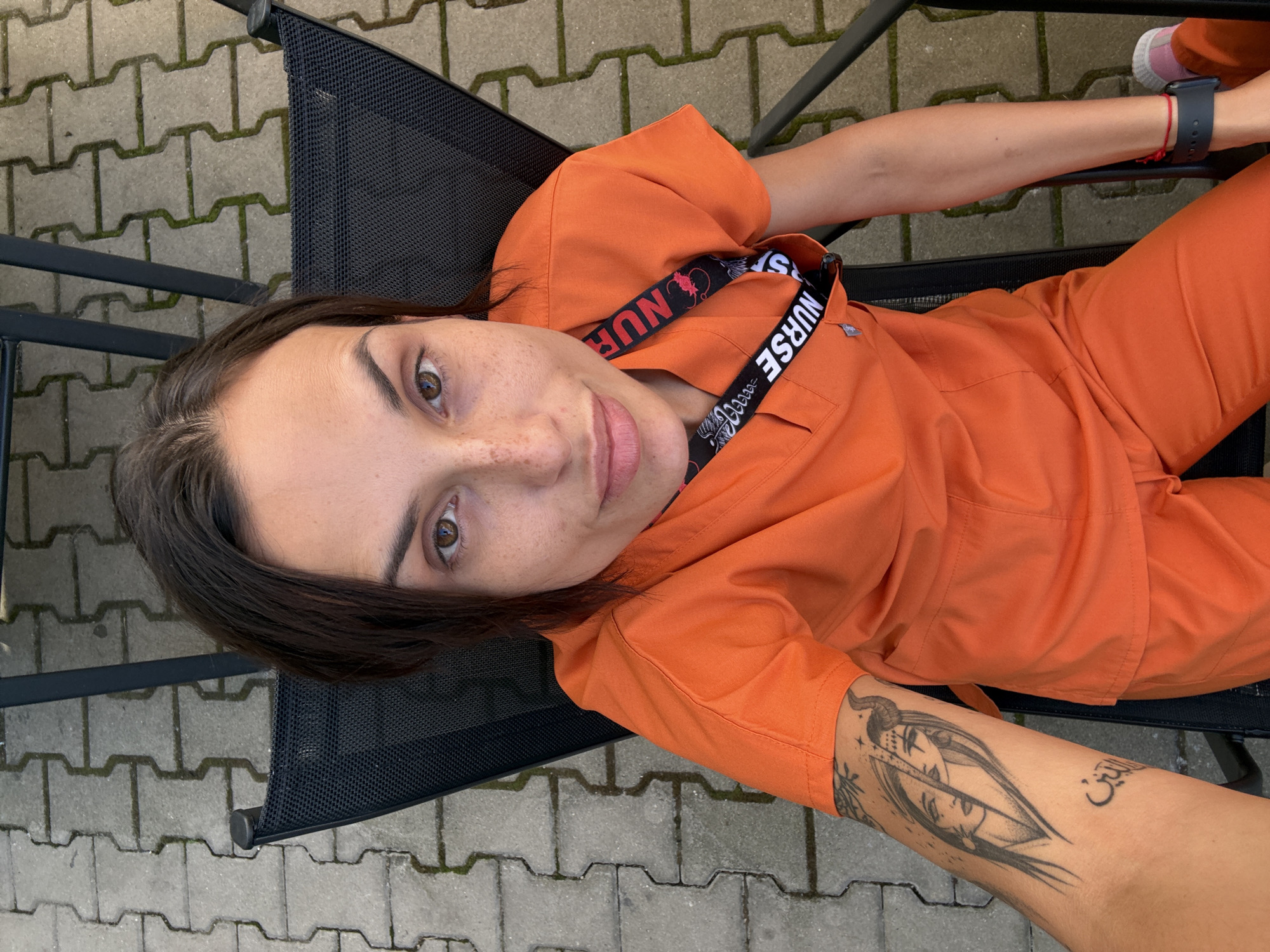 A high-angle, medium shot captures a woman with dark hair and freckles, wearing orange scrubs, sitting outdoors on a black mesh chair. She is looking directly at the camera with a slight smile. A black lanyard with "NURSE" printed on it is visible around her neck. Her left arm, resting on her leg, displays a tattoo of two faces and Arabic script. The background consists of a paved patio area with interlocking concrete slabs and green moss. The setting is Pleven, Bulgaria.