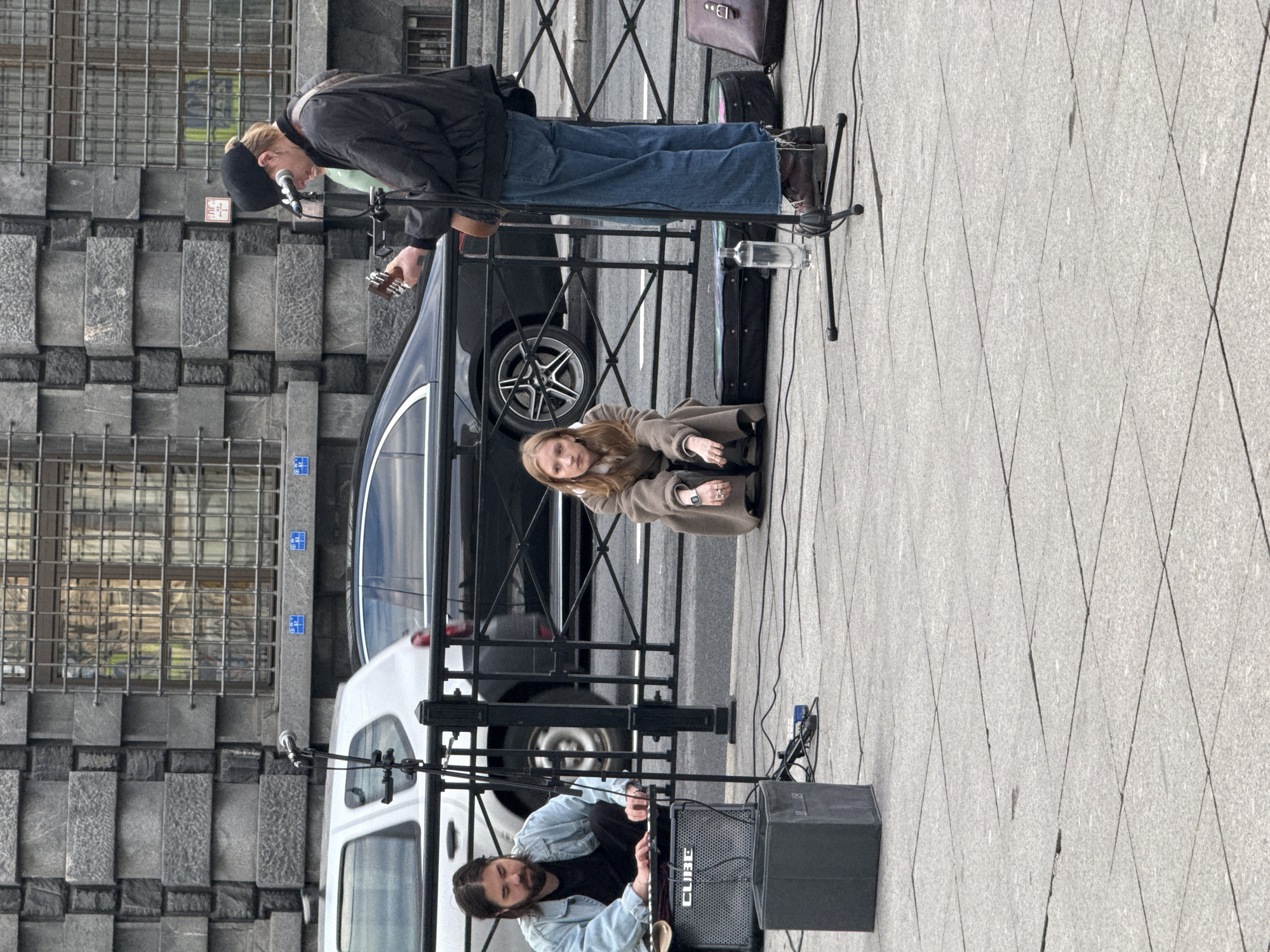 The image shows a street performance in Saint Petersburg, Russia. The scene features three people: a man playing a guitar and singing into a microphone, a woman sitting on the ground nearby, and another man kneeling by a speaker. The man playing the guitar is wearing a black jacket and blue jeans. The woman is dressed in a brown coat and appears to be listening to the music. The man by the speaker is wearing a denim jacket and is interacting with the equipment.
The setting is a city sidewalk with a stone building facade in the background and a black car parked behind a metal fence. There are microphones, a guitar, and a speaker visible, indicating a musical performance. The weather appears overcast. The time of day is indeterminate, but likely daytime.
Visible text includes "CUBE" on the speaker. The overall mood seems casual, with the performers and audience member engaged in the music. The architecture and the presence of a car suggest an urban environment.