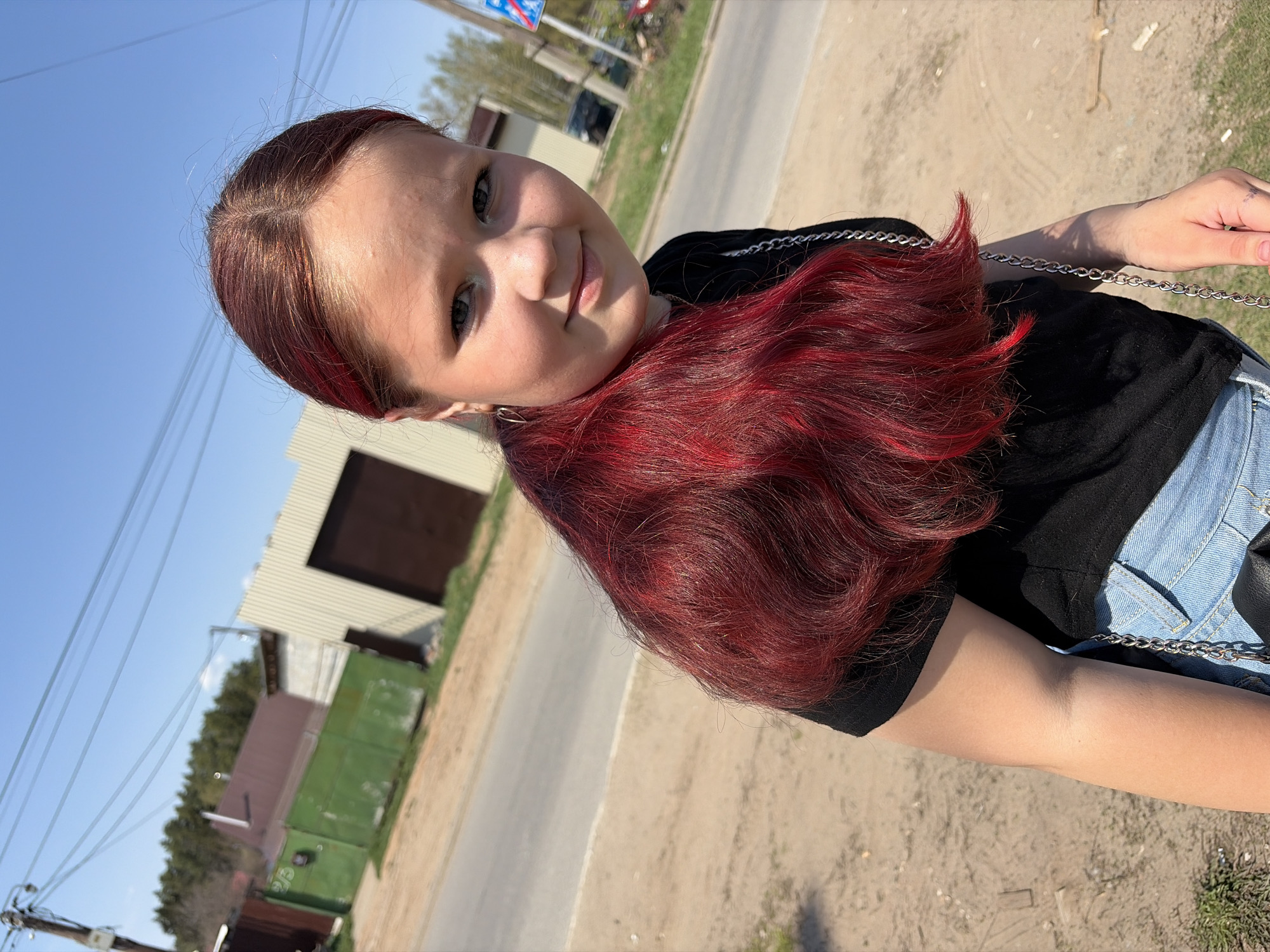 A young, fair-skinned person with long, dark red hair and wearing a black top and blue jeans stands outdoors. They are likely positioned near a road or sidewalk. A building with a brown door and a green structure is visible in the background. The location is in Chaikovsky, Russia.