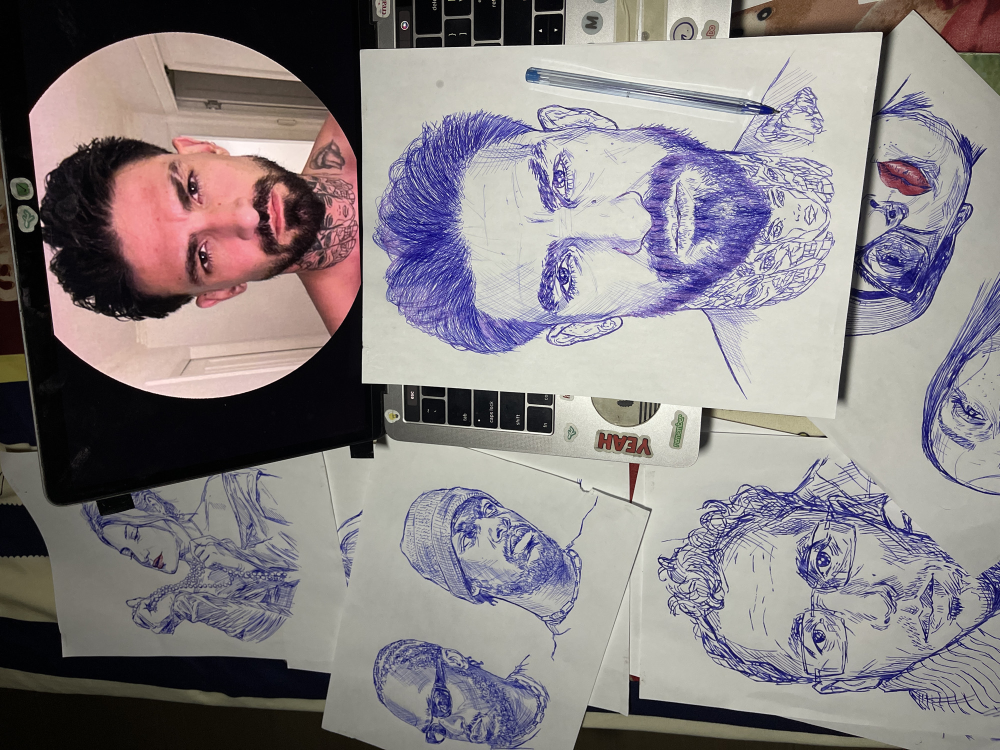 The image depicts a collection of pen and ink drawings of portraits, predominantly in blue ink with some red accents. The drawings are laid out on a surface, possibly a desk or table, which is covered with a striped cloth. A laptop is partially visible, with a circular photo displayed on its screen showing a man with dark hair, a beard, and tattoos on his neck.
The drawings include:
- A woman holding a pearl necklace, with red lipstick.
- Two men, one wearing sunglasses and a chain, the other wearing a beanie and a chain.
- A close-up portrait of a man's face, with detailed hair and beard.
- A portrait of a man wearing glasses.
- A partial drawing of a woman's face with striking red lips.
A blue ballpoint pen lies on top of one of the drawings. Some stickers, including one that says "YEAH" and another that says "remember", are visible on the laptop. The overall setting appears to be an artist's workspace, with various sketches and art supplies. The image does not contain any elements that indicate it is located in the city of Jos, Nigeria.