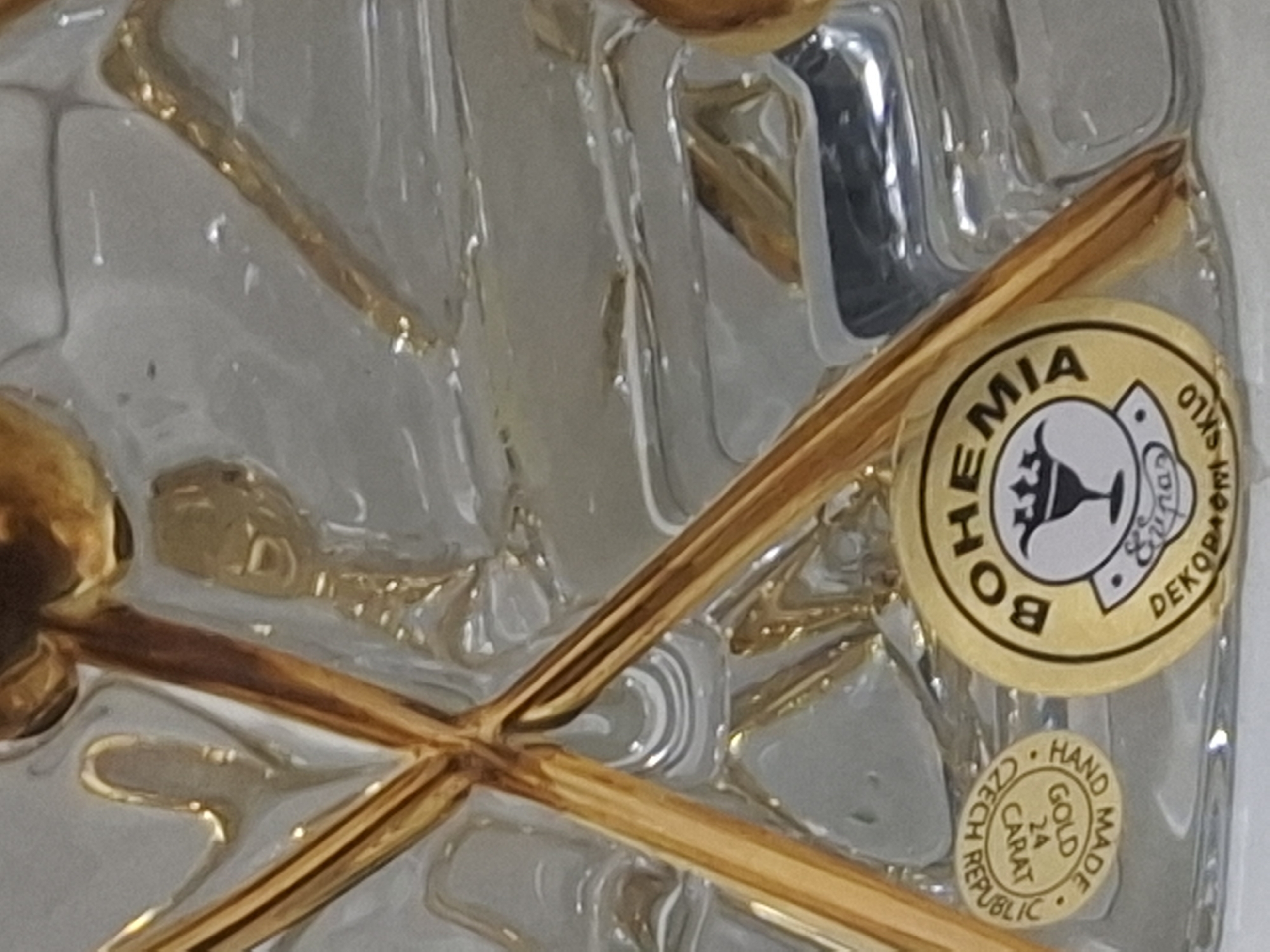 The image is a close-up shot of a crystal vase with gold-colored accents. Two round, gold-colored stickers are visible on the vase. The sticker on the left is smaller and reads "HAND MADE CZECH REPUBLIC 24 CARAT GOLD". The larger sticker on the right has "BOHEMIA" at the top and appears to be a manufacturer's or brand label. The vase itself has a faceted design with diagonal gold elements crossing over the crystal. The background is out of focus, but appears to be a reflective surface, possibly a table. There are no people or any discernible environmental features that would indicate a specific location within Almaty, Kazakhstan.