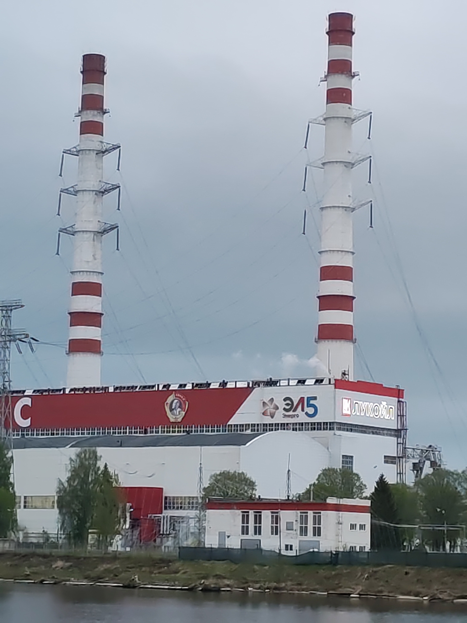 The image shows a large industrial complex, likely a power plant, with two tall chimneys prominently featuring red and white stripes. The chimneys have electrical structures running up the sides. The main building is painted white with a red band at the top. On the red band are the large letter "C", a symbol with what appears to be a person's face, the symbol "ЭЛ5", and the word "ЛУКОЙЛ" in Cyrillic script. The location is adjacent to a body of water, suggesting a riverside or coastal location. Smaller white buildings with red roofs are situated in front of the main structure. Trees are visible along the waterfront, with overcast skies in the background. There are no visible people or interactions in the scene.