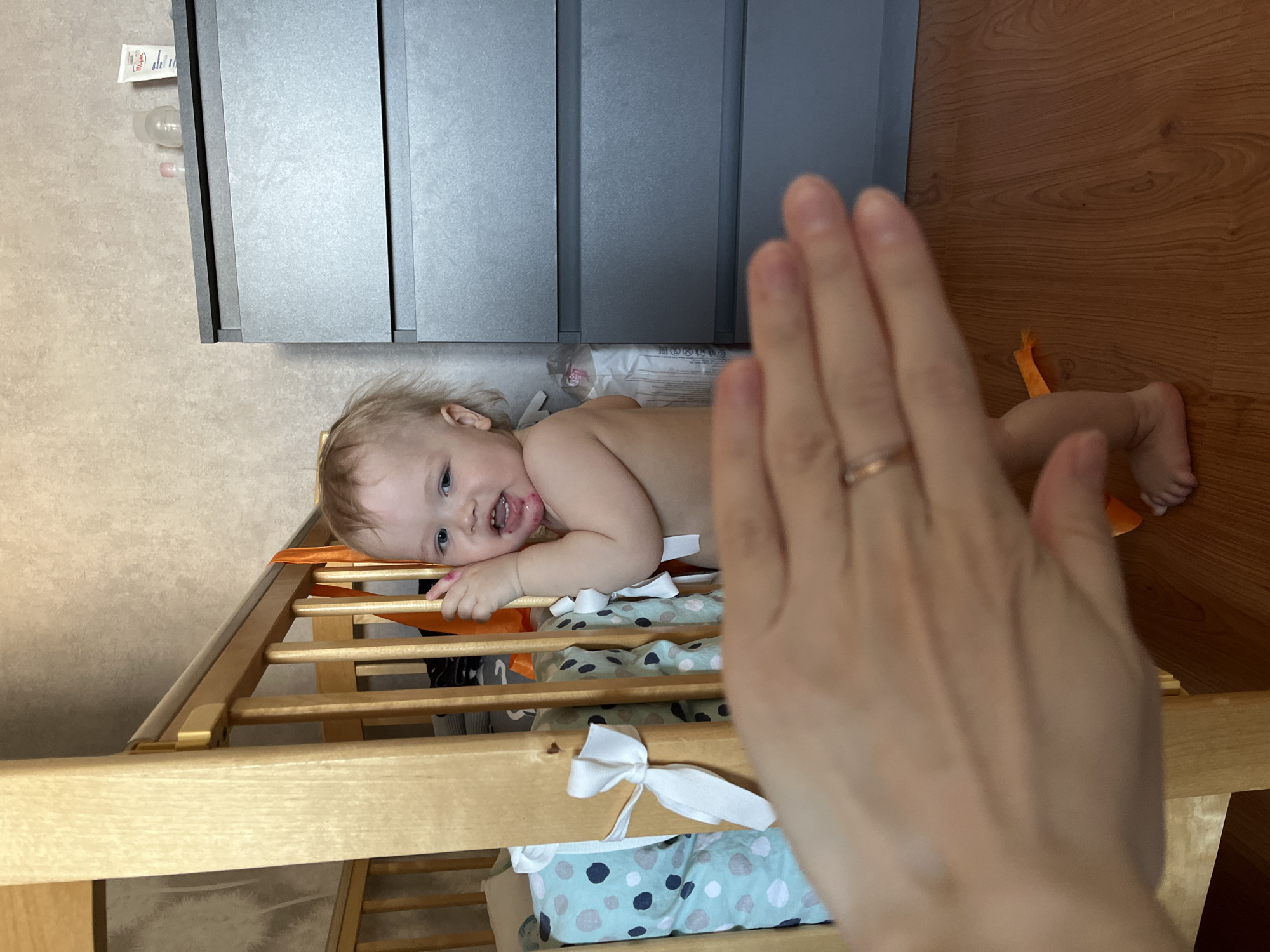 A baby is lying on a changing table in what appears to be a domestic setting, perhaps in Chaikovsky, Russia. The baby, who has light hair and blue eyes, is on their stomach, facing the viewer. The changing table is wooden and has a patterned changing pad on top. A hand is raised in the foreground, partially obscuring the scene. There are cabinets above the changing table and wooden flooring visible in the background.